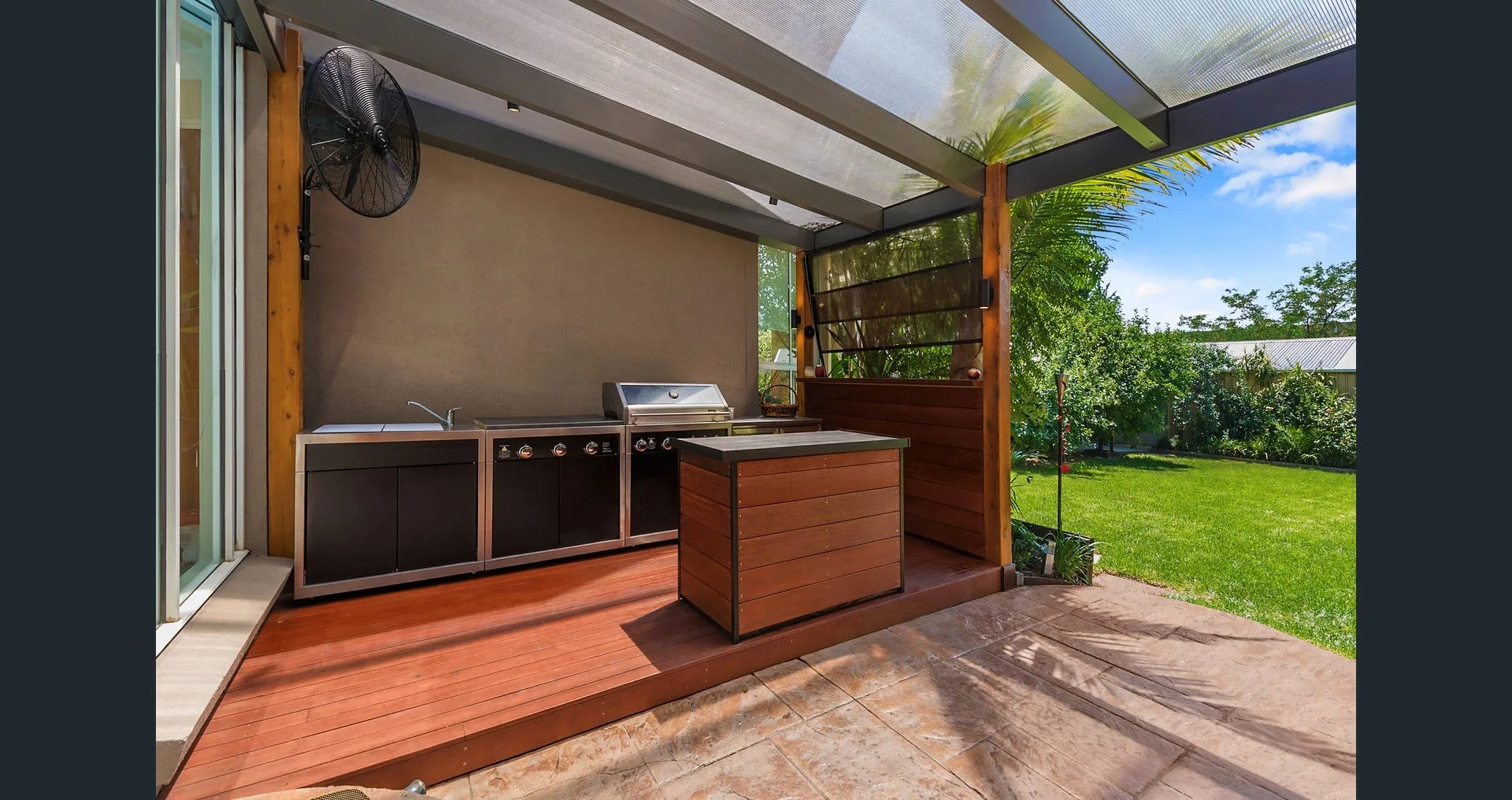 Outdoor kitchen area with a stainless steel grill, small sink, and cabinetry, under a metal and glass covered patio with a view of a green backyard with trees and a blue sky.