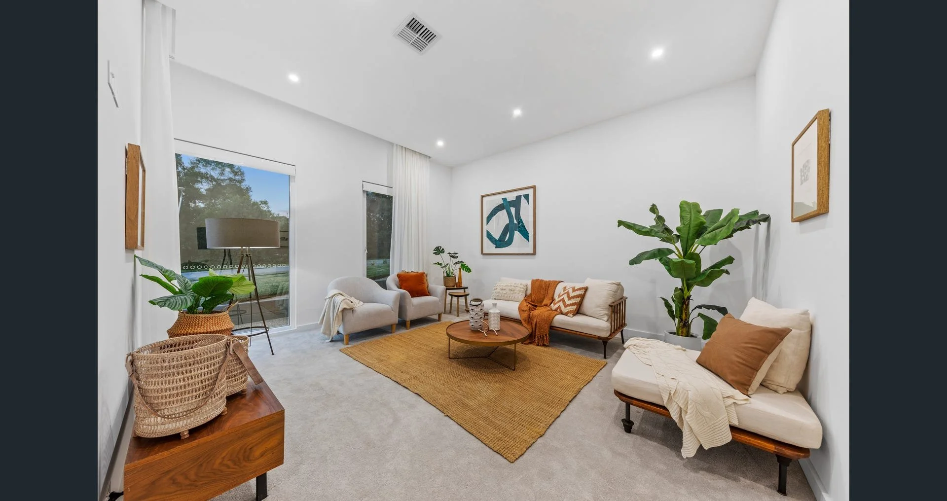 Living room with white walls, multiple windows, and a variety of seating including a beige armchair, a white couch, and a wooden bench with a pillow, beige rug, indoor plants, artwork, and a sliding glass door leading to an outdoor area.