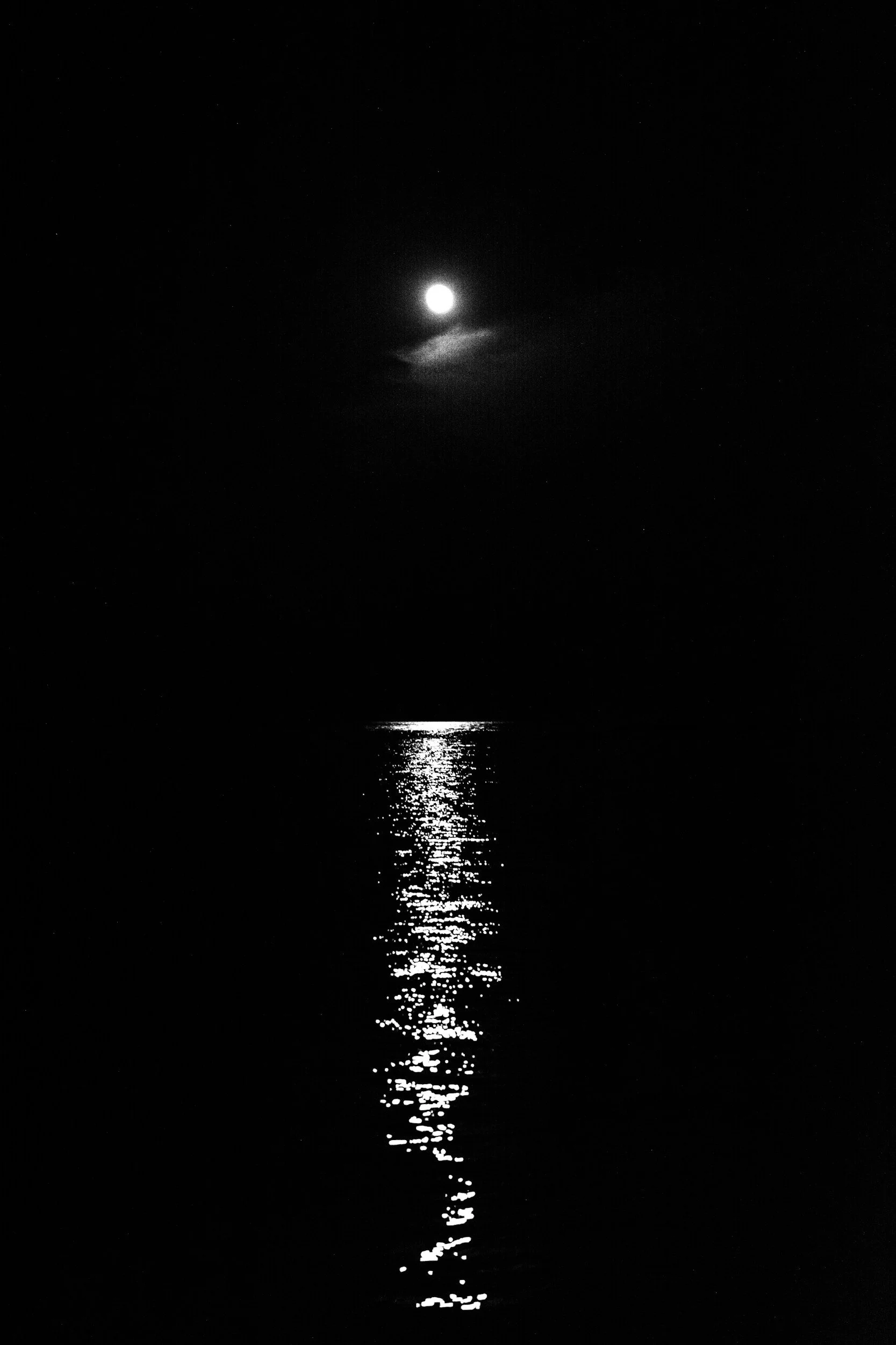Black and white photo of the moon reflecting on the ocean, with a small cloud near the moon and stars visible in the night sky.