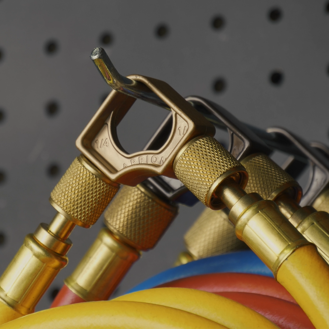 Close-up of brass high-pressure hydraulic fittings with yellow, red, and blue hoses attached, placed on a grey pegboard background.