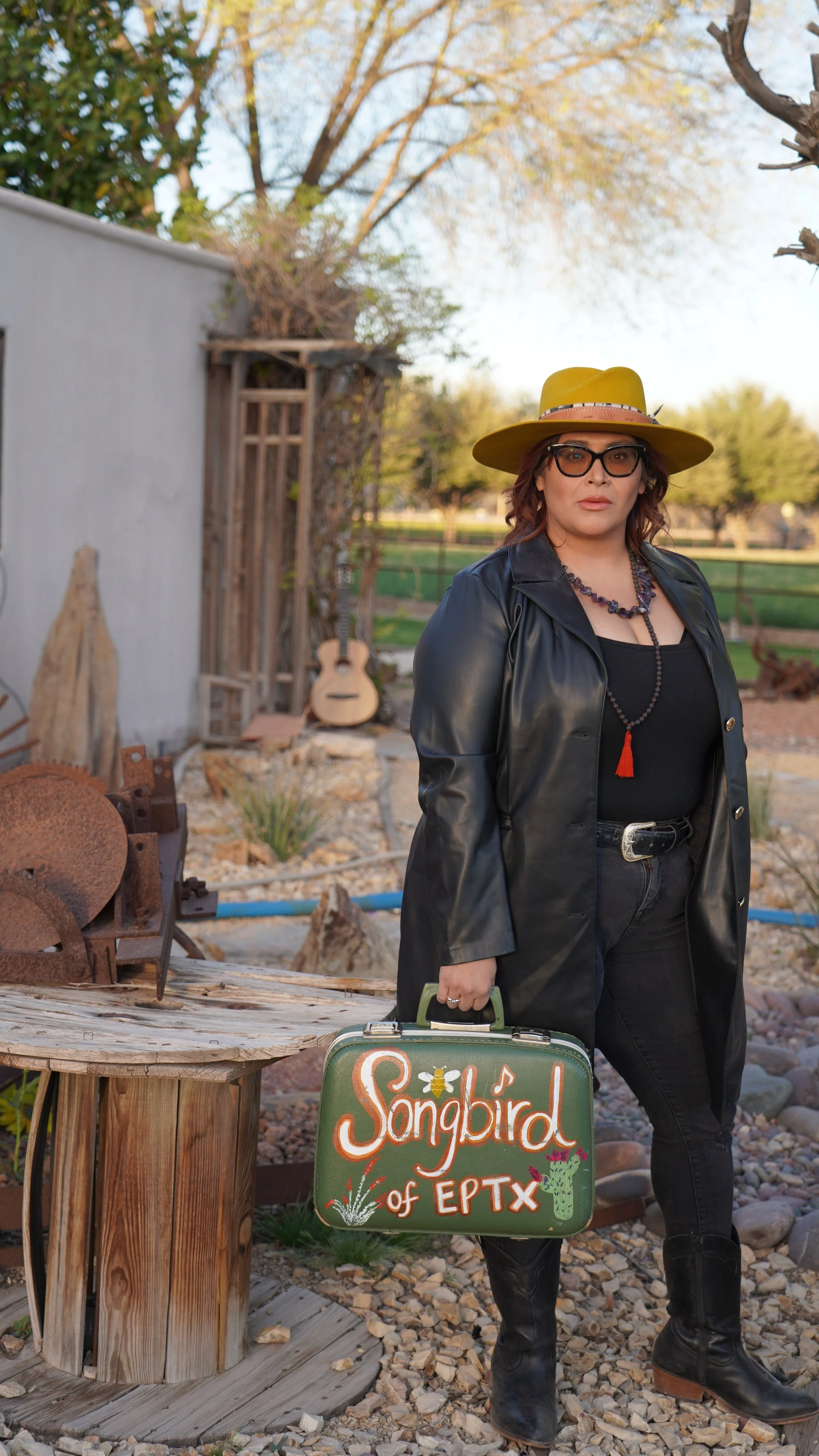 A woman with glasses and wavy hair wearing a yellow hat, black leather jacket, black top, and black pants with black boots, standing outdoors and holding a vintage green suitcase with the words "Songbird of EPTX" painted on it, in a rustic outdoor setting with trees, a guitar, and various decorative items.