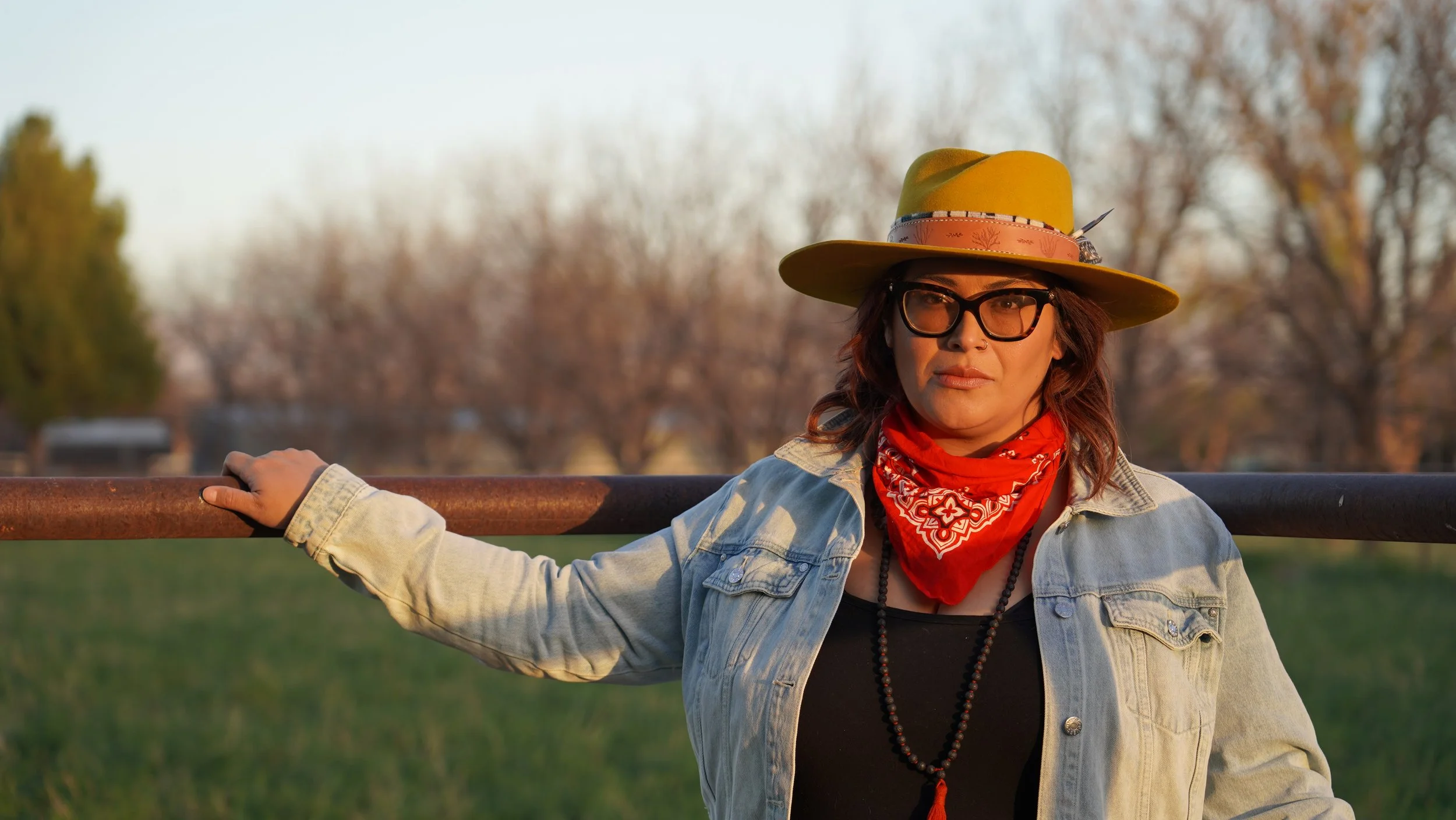 Woman with glasses and a red bandana around her neck, wearing a yellow hat and denim jacket, standing outdoors at sunset, holding a wooden rail, with trees in the background.