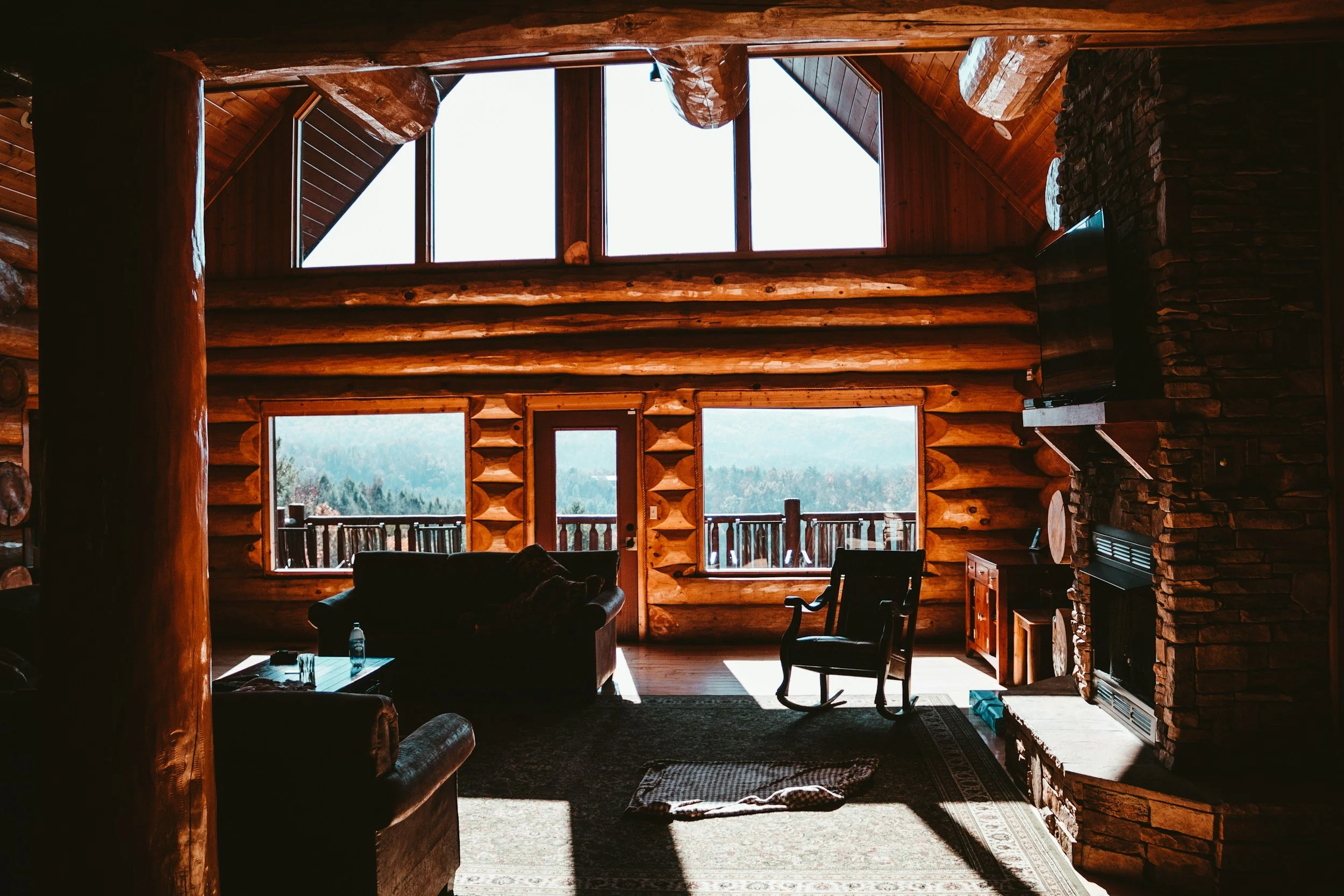 Interior of a rustic cabin living room with large windows showing a mountain view, wood log walls, a stone fireplace, and furniture including a rocking chair, sofa, and coffee table.