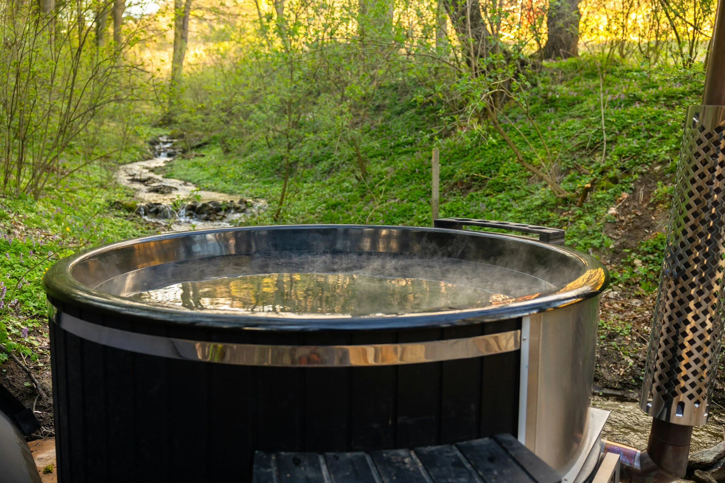 Hot tub outdoors in a lush green forested area with a small stream in the background