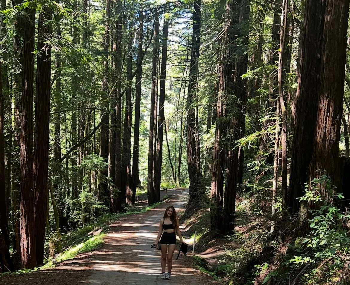A young woman walks along a dirt trail through a dense forest of tall trees. She is holding a camera in one hand and a jacket in the other, smiling as she enjoys her hike.