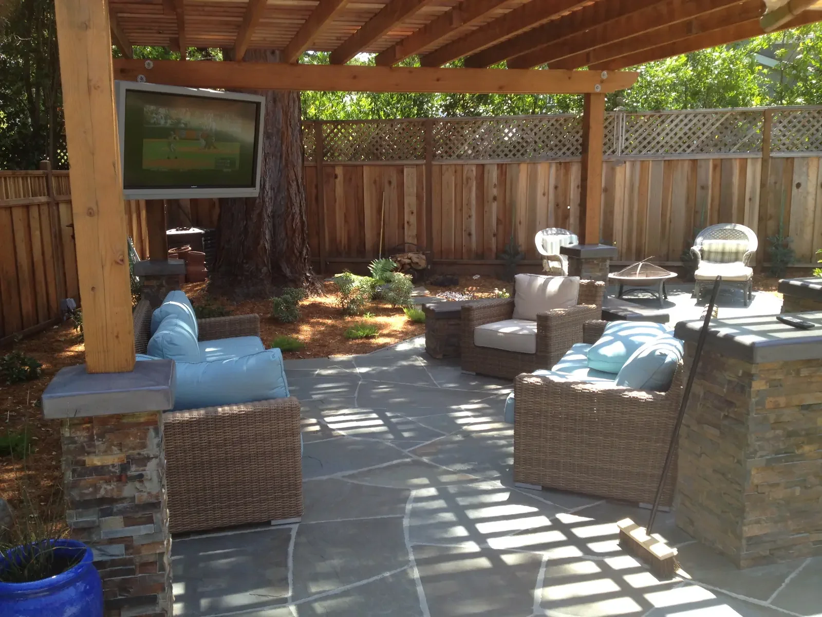 Outdoor patio area with wicker chairs and sofas, a television mounted to a wooden post under a pergola, and a wood privacy fence. Shaded and sunlit areas with trees in the background.