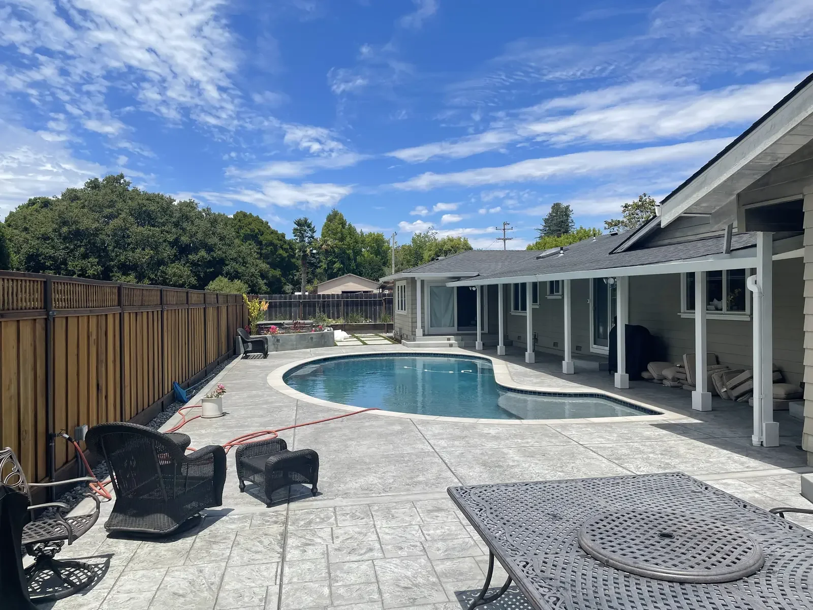 A backyard with a kidney-shaped swimming pool surrounded by a stone deck, enclosed by a wooden fence. There are black outdoor chairs, a table, and potted plants. The house has a covered patio area with stacked chairs and a lawn in the background with trees and a blue sky with clouds.
