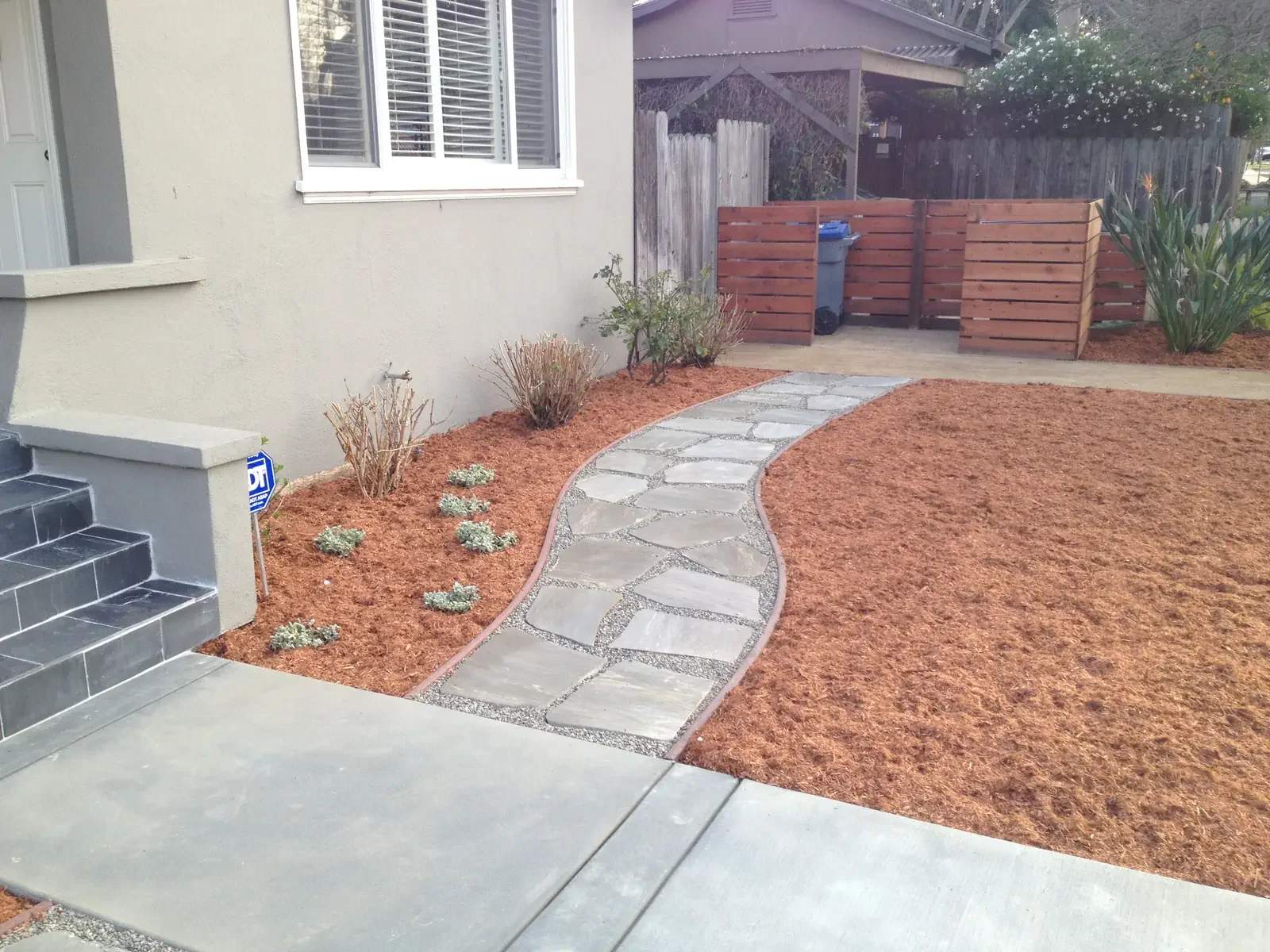 A concrete sidewalk with a curved stone pathway leading to a wooden gate in a fenced backyard. The yard has red mulch and small plants along the house wall, with a trash bin near the gate.