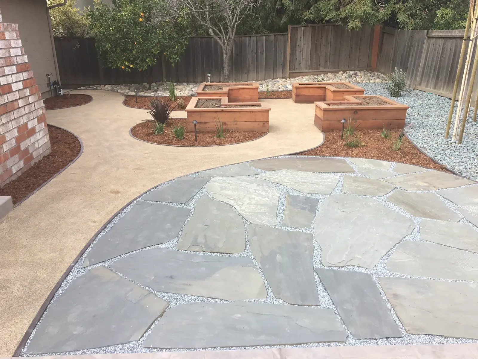 A backyard with a stone patio, a concrete pathway, and raised wooden garden beds, surrounded by a wooden fence and trees.