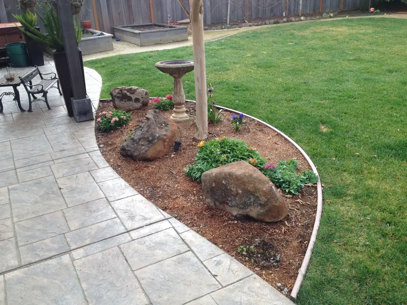 A backyard garden with a landscaped flower bed containing rocks, flowers, a birdbath, and a tree, bordered by a lawn and a stone patio.