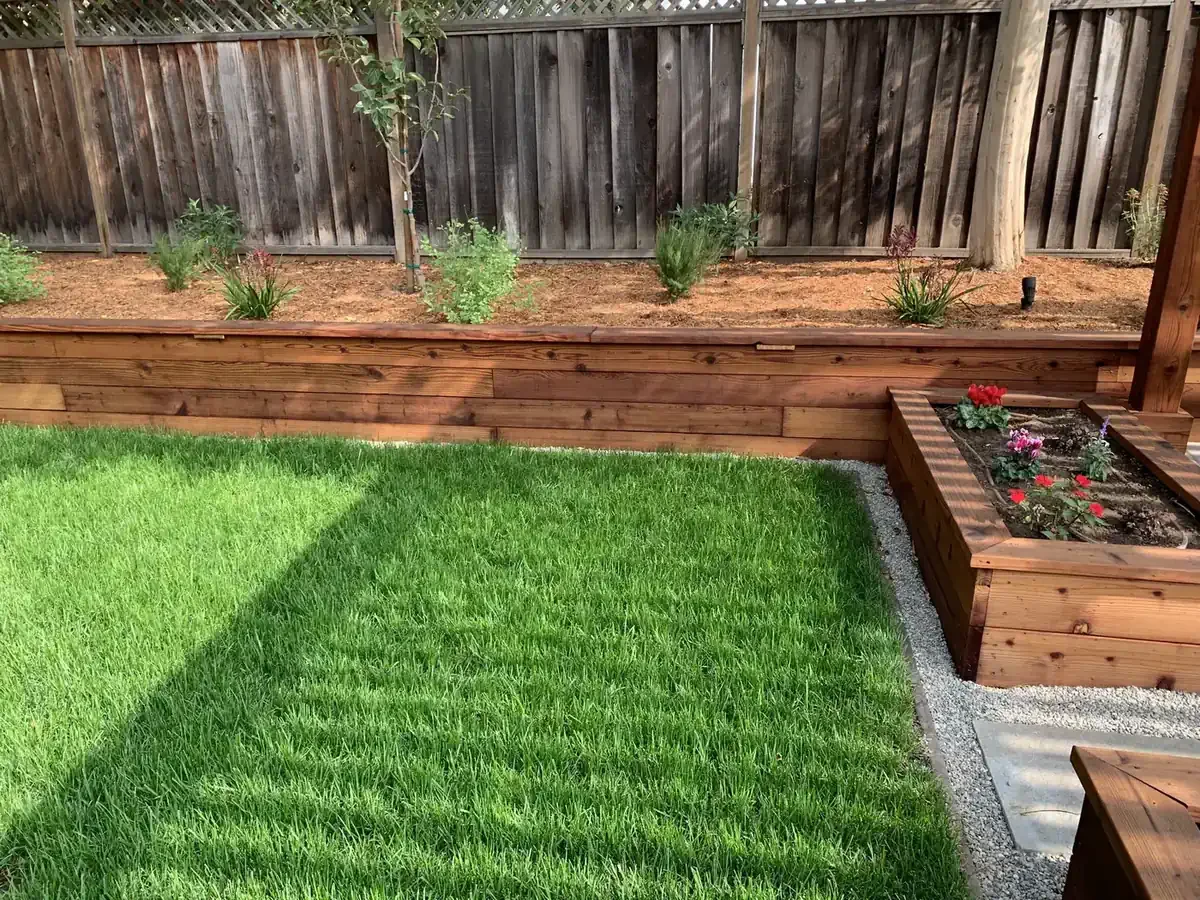 A backyard with a well-maintained green lawn, a wooden planter box with colorful flowers, and a raised garden bed with small plants, all enclosed by a wooden fence.