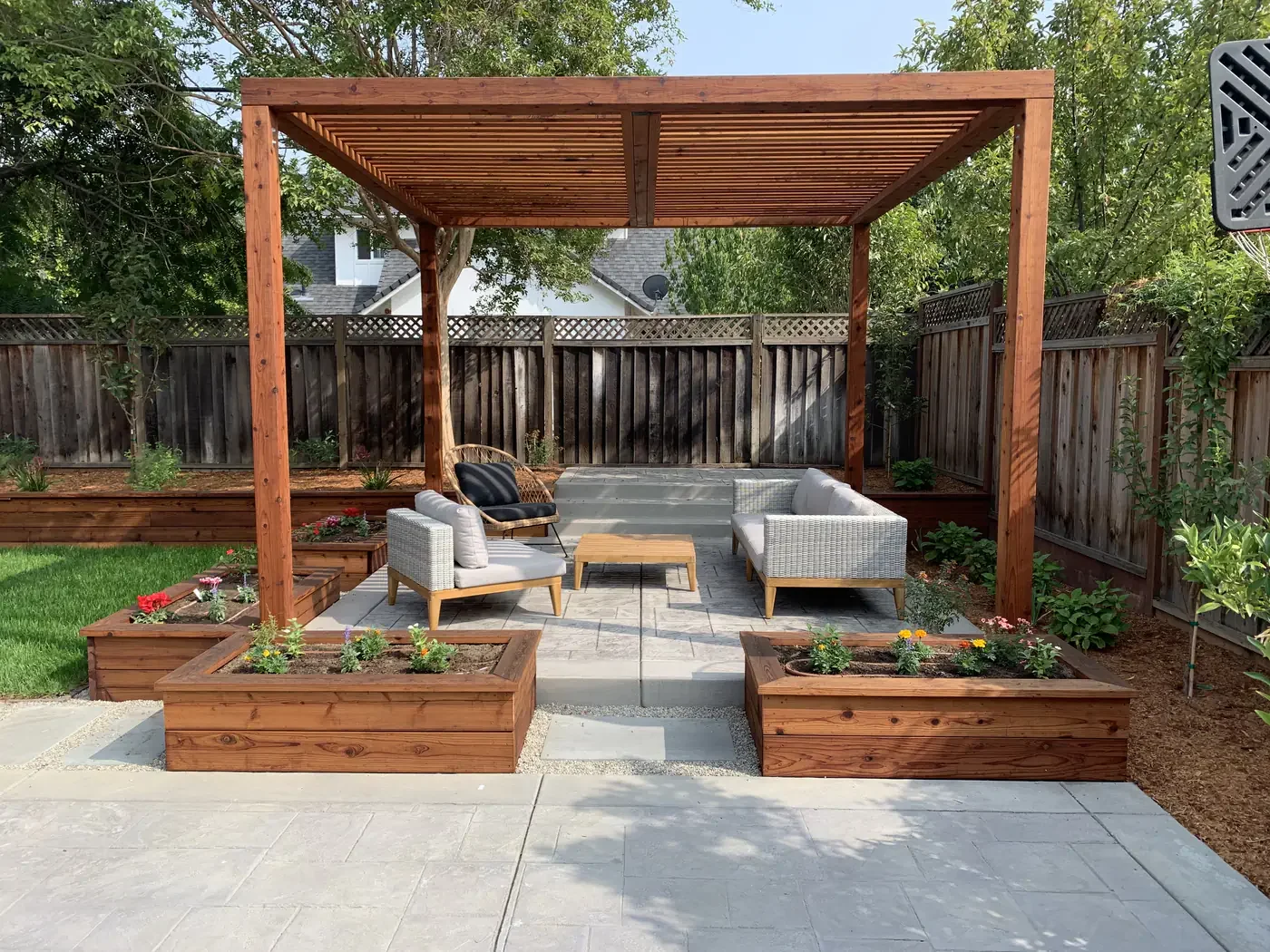 Backyard patio with wooden pergola, outdoor seating, flower beds, and a privacy fence.
