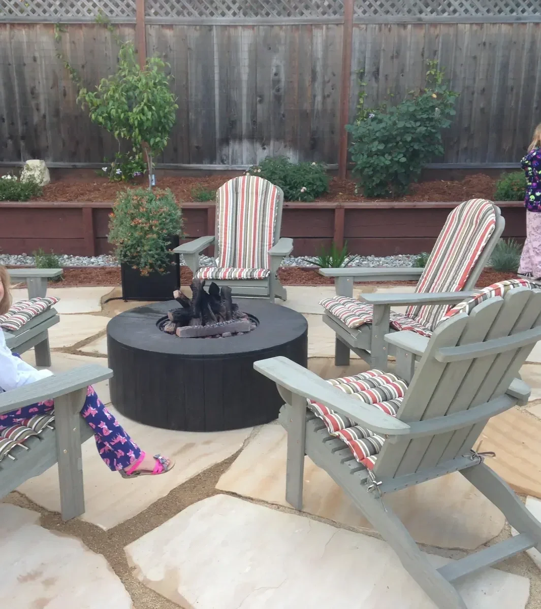 Backyard patio with four Adirondack chairs with striped cushions, a fire pit in the center, potted plant, and a raised garden bed with bushes and small trees against a wooden fence.