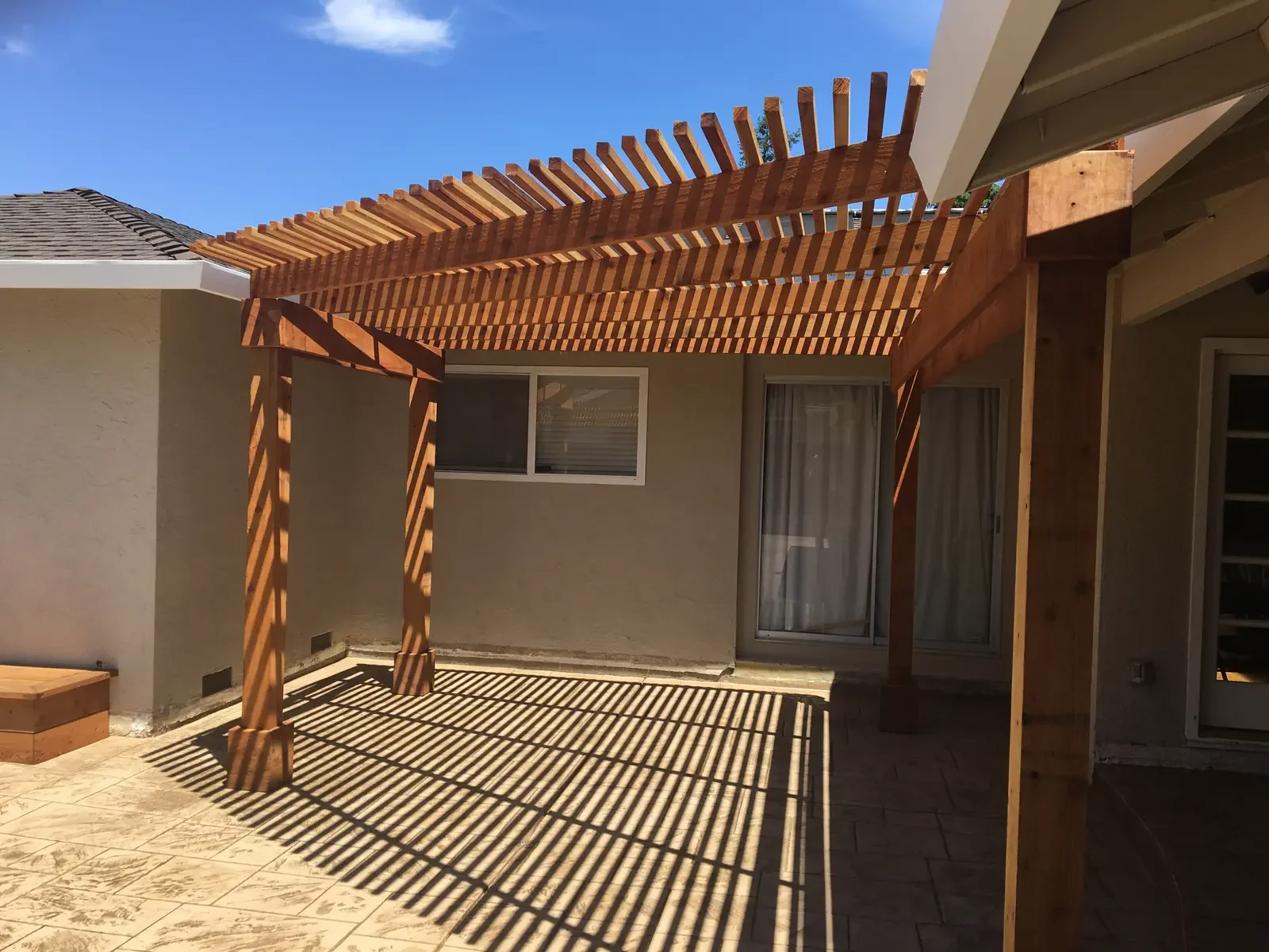 Wooden pergola on a patio, casting striped shadows on the concrete floor of a backyard.