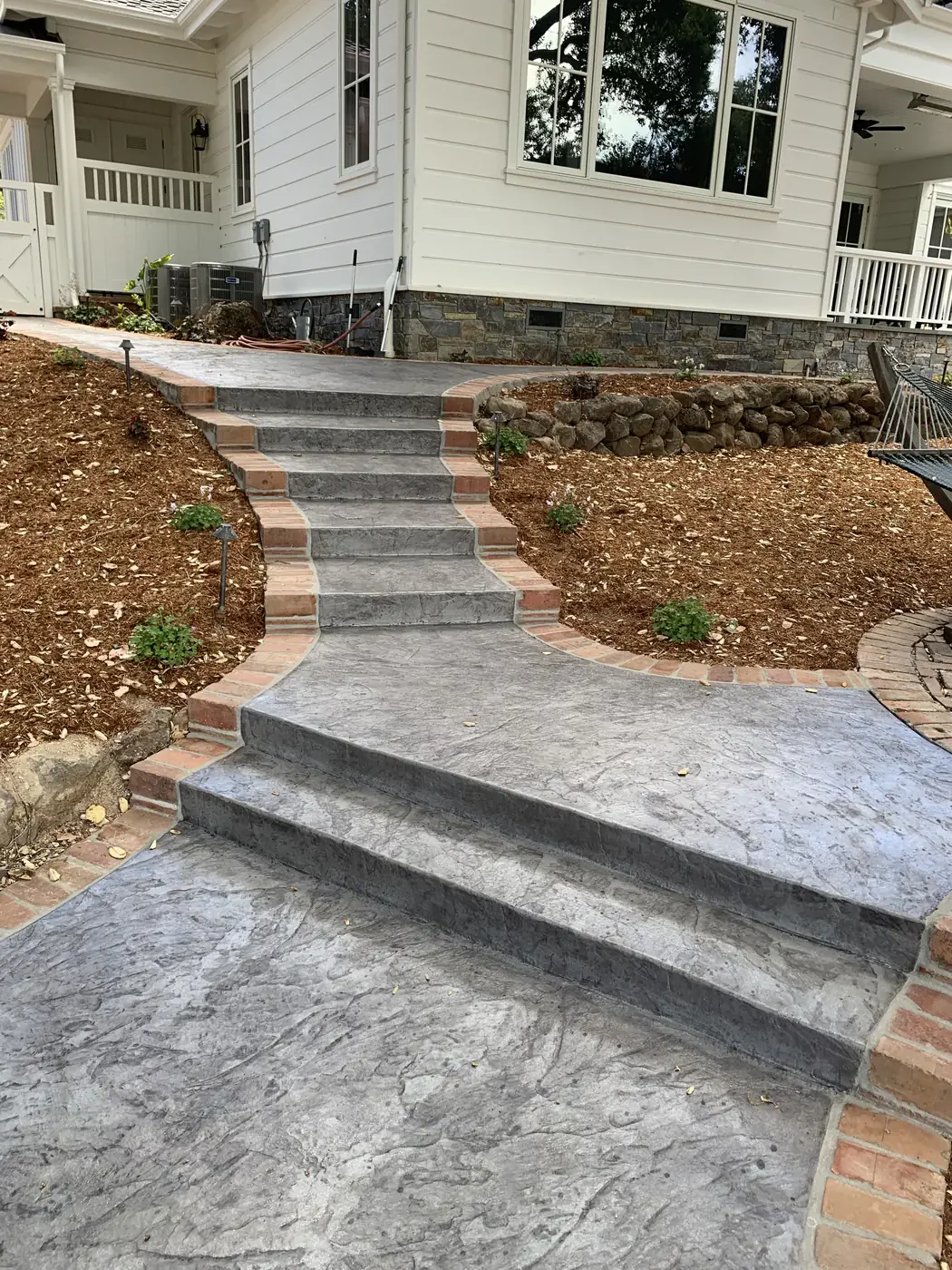 A concrete pathway with stairs leads from the yard to the front porch of a white house with large windows and a small balcony. The pathway is bordered by bricks and surrounded by brown dirt and small plants.