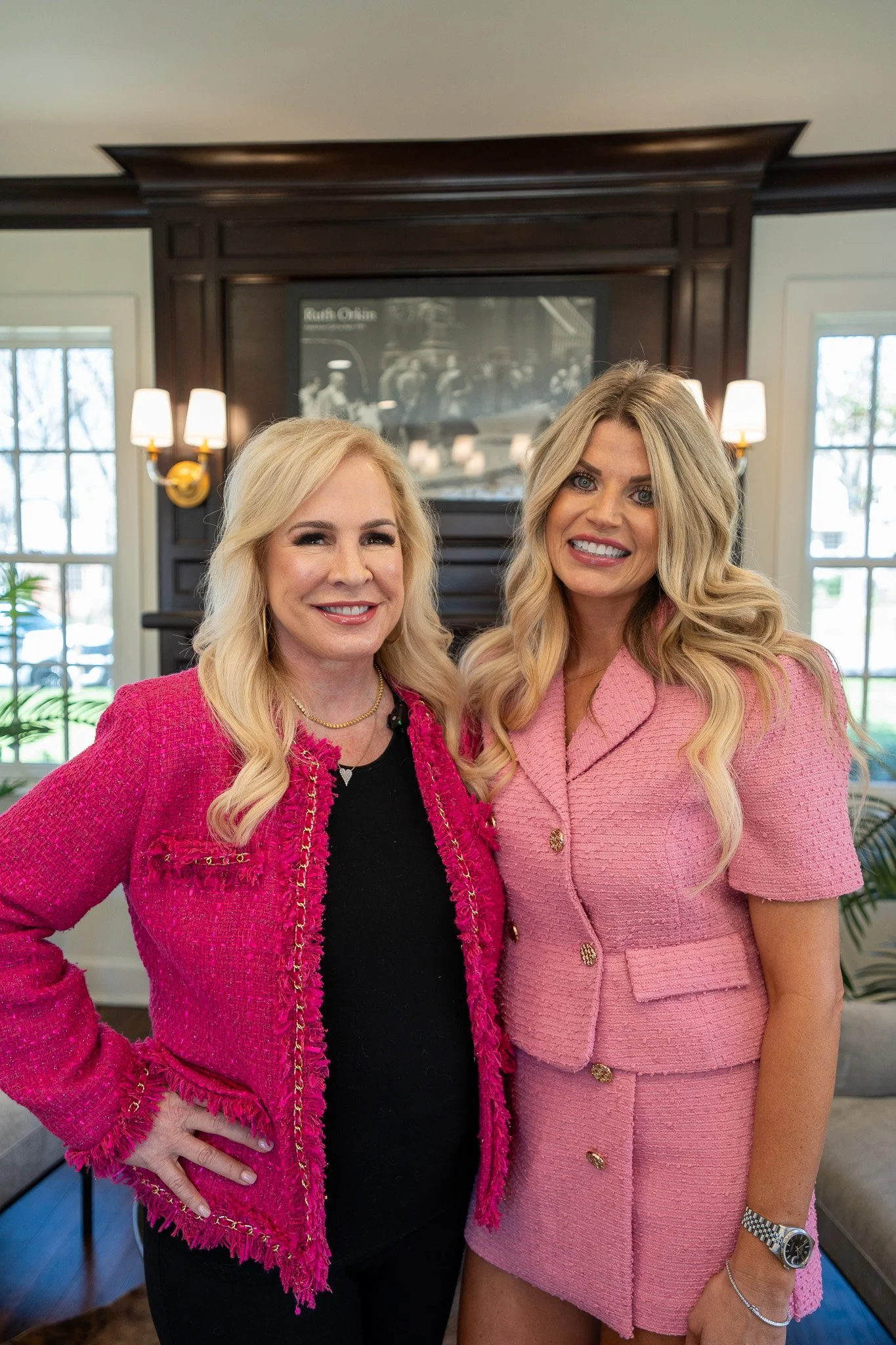 Two women with blonde hair smiling and standing indoors near a fireplace, wearing pink and black clothing.