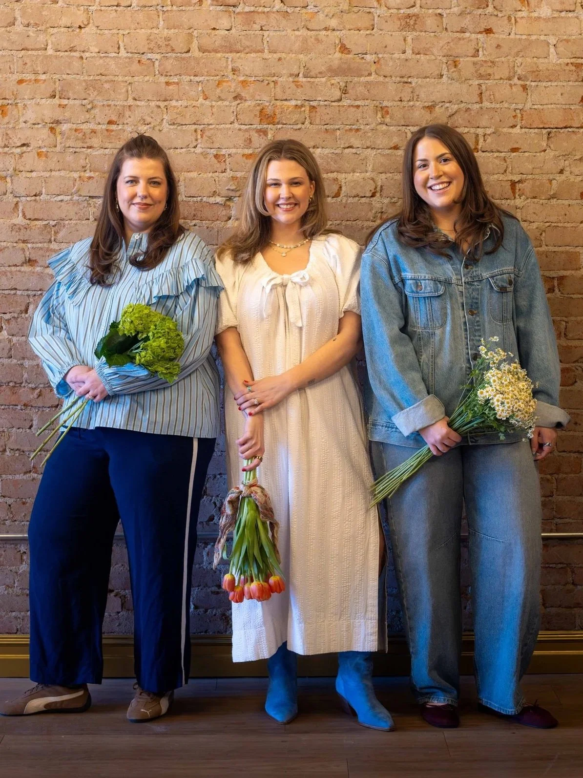 Three women standing against a brick wall, each holding a bouquet of flowers. The woman on the left has brown hair and wears a striped blouse with ruffled sleeves, holding green flowers. The woman in the center has light brown hair and wears a white dress, holding a bouquet of pink tulips. The woman on the right has dark brown hair and wears a denim jacket and jeans, holding a bouquet of small white flowers.