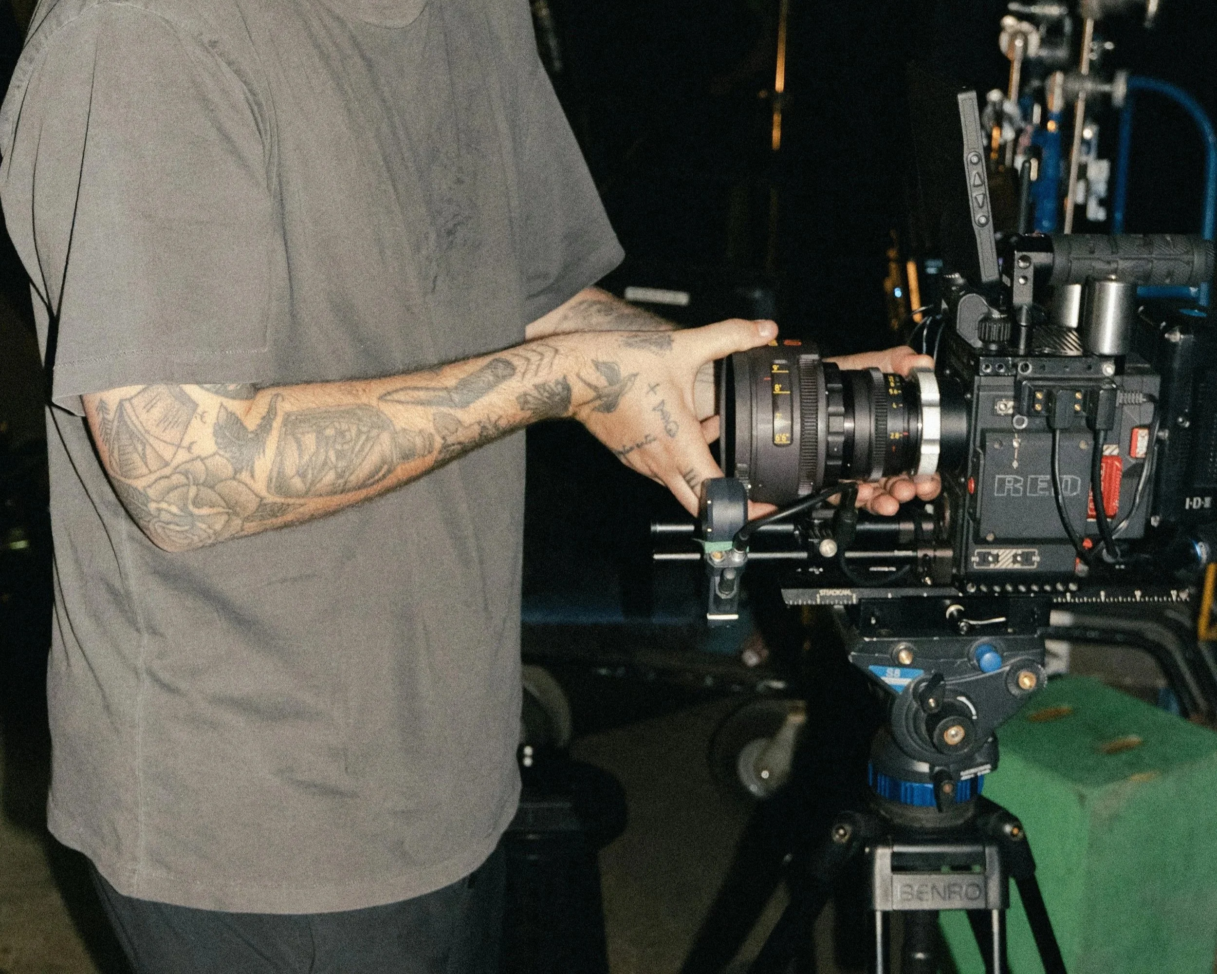 A person with tattooed arm operating a professional movie camera on a tripod in a dimly lit environment.