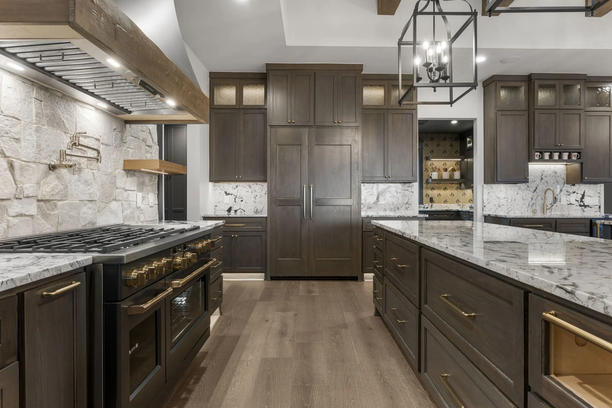 A modern kitchen with dark wooden cabinets, a marble countertop, a built-in oven with brass knobs, a stone wall with brass faucets, and a wooden ceiling beam with a lighting fixture.