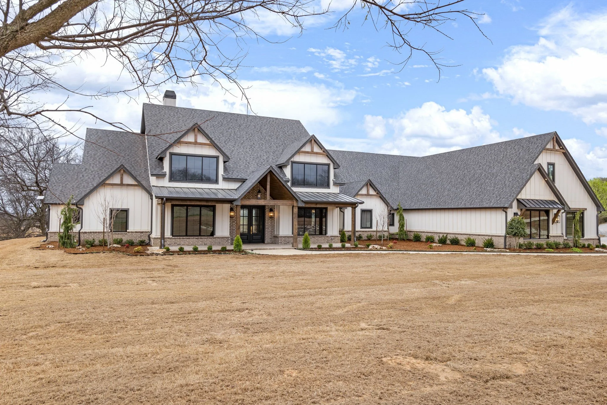 Large modern house with a gray roof, white exterior, and black doors, surrounded by a landscaped yard with small bushes and young trees.