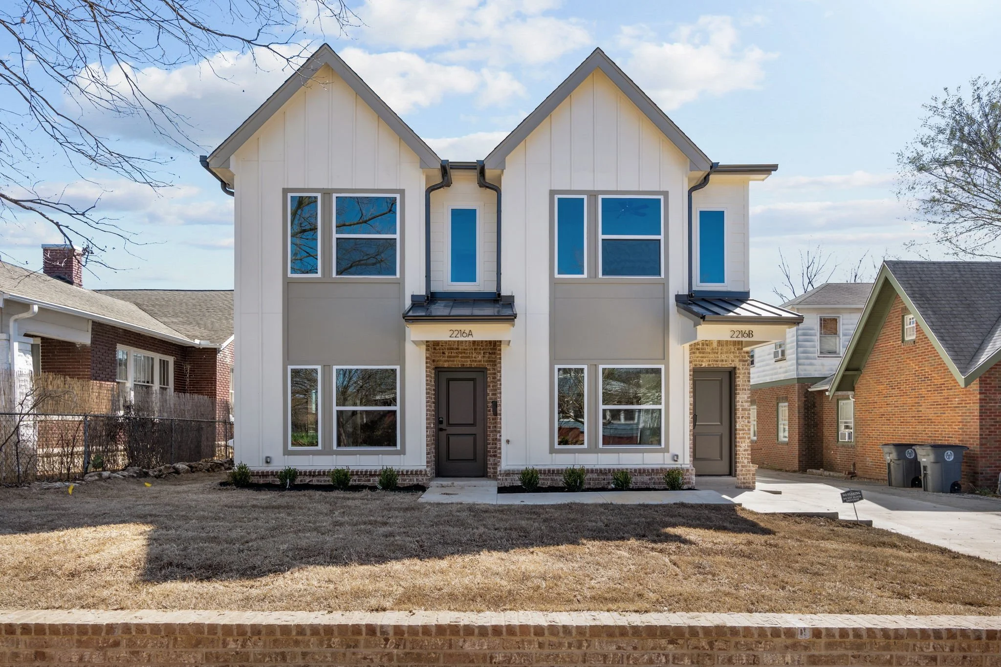 A modern duplex home with white siding, brown doors, and large blue-tinted windows, surrounded by a small lawn and neighboring houses, under a partly cloudy sky.