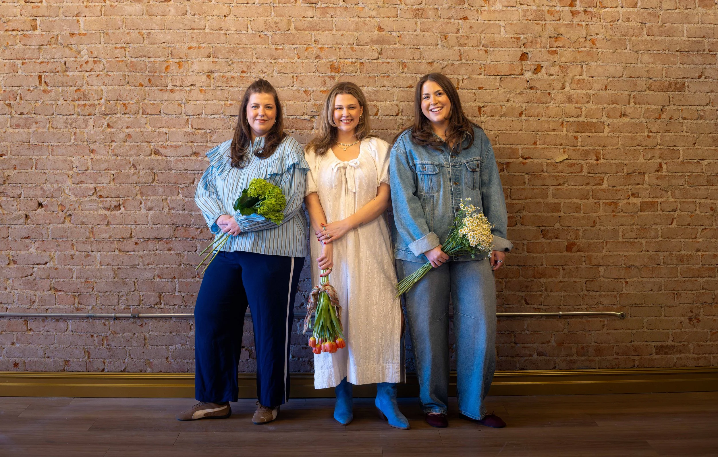 Three women standing against a brick wall, each holding a bouquet of flowers, smiling at the camera.