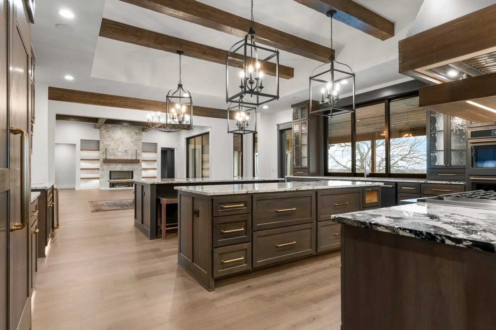 Modern kitchen with dark wooden cabinets, marble countertops, and a kitchen island. Hanging chandeliers with black frames and candles. Large windows revealing outdoor trees, and a white living room with fireplace in the background.