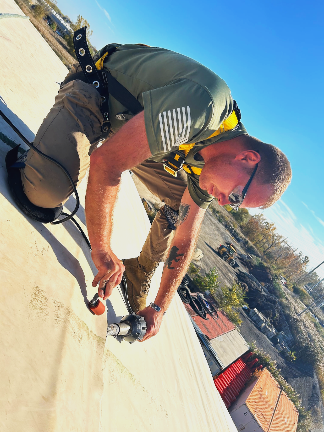 A construction worker with sunglasses, a green shirt, and khaki pants kneeling on a flat surface while using a power tool. The background features an outdoor setting with trees, buildings, and parked vehicles under a clear blue sky.