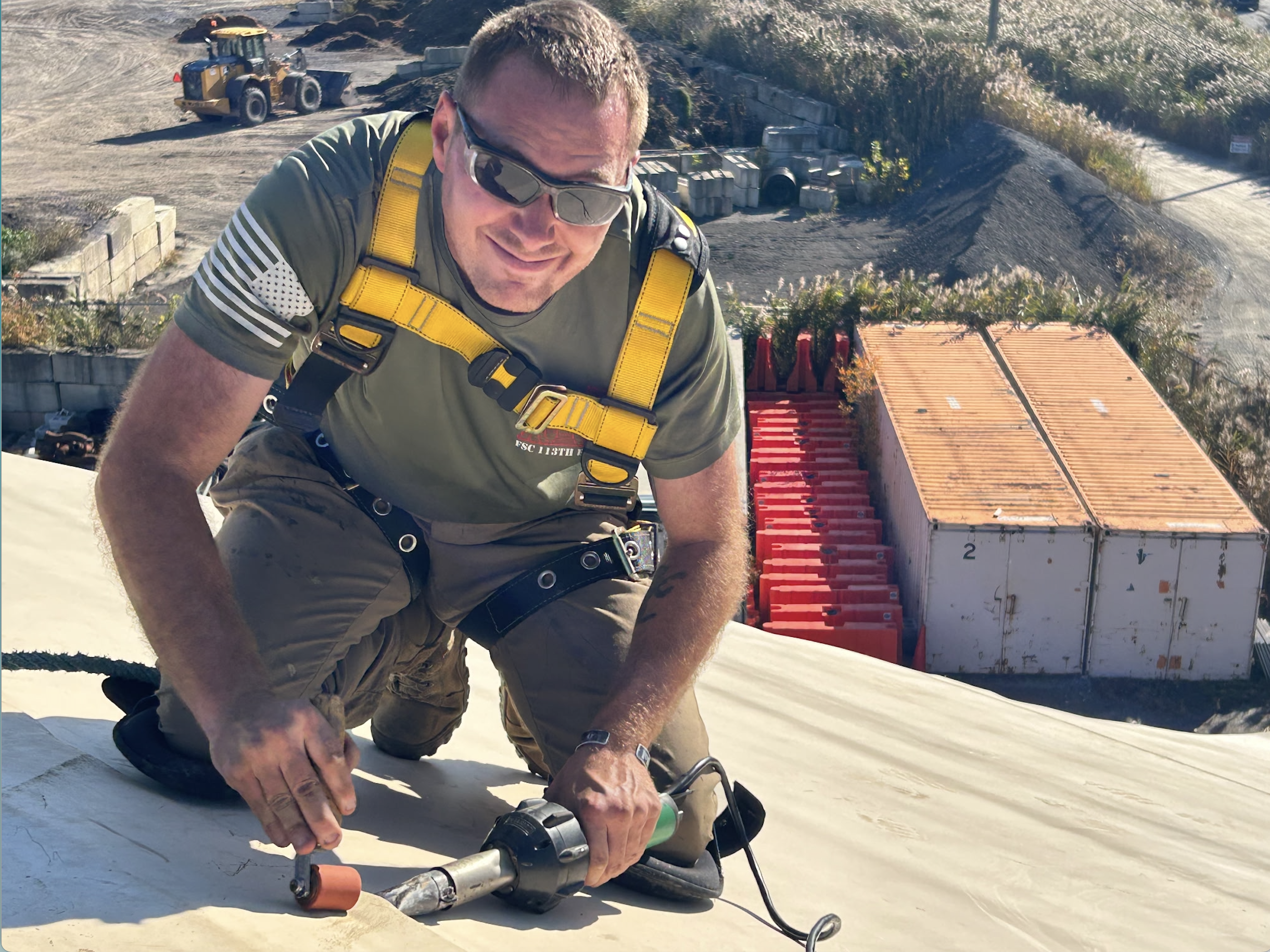 A man in safety glasses and a green shirt with yellow suspenders kneels on a roof, using a power saw to cut the roofing material. Construction materials and equipment are visible in the background, including stacked shipping containers, a loader, and a pile of dirt.