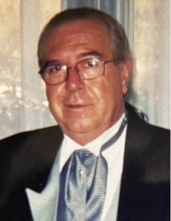 A man with glasses wearing formal attire, including a tuxedo with a bow tie, in front of a window with curtains.