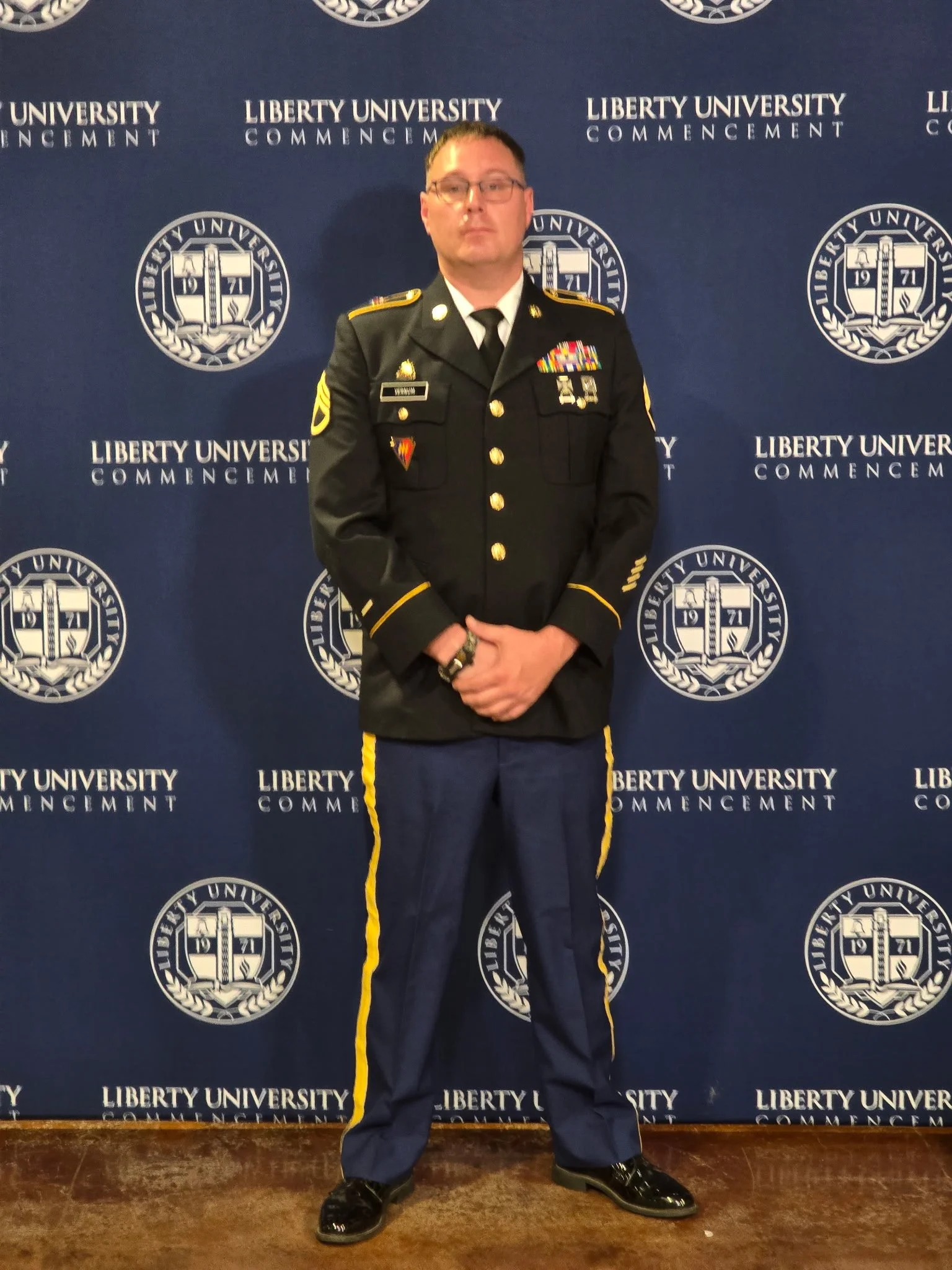A man in a military dress uniform standing in front of a blue backdrop with Liberty University logos, at a graduation or commencement event.
