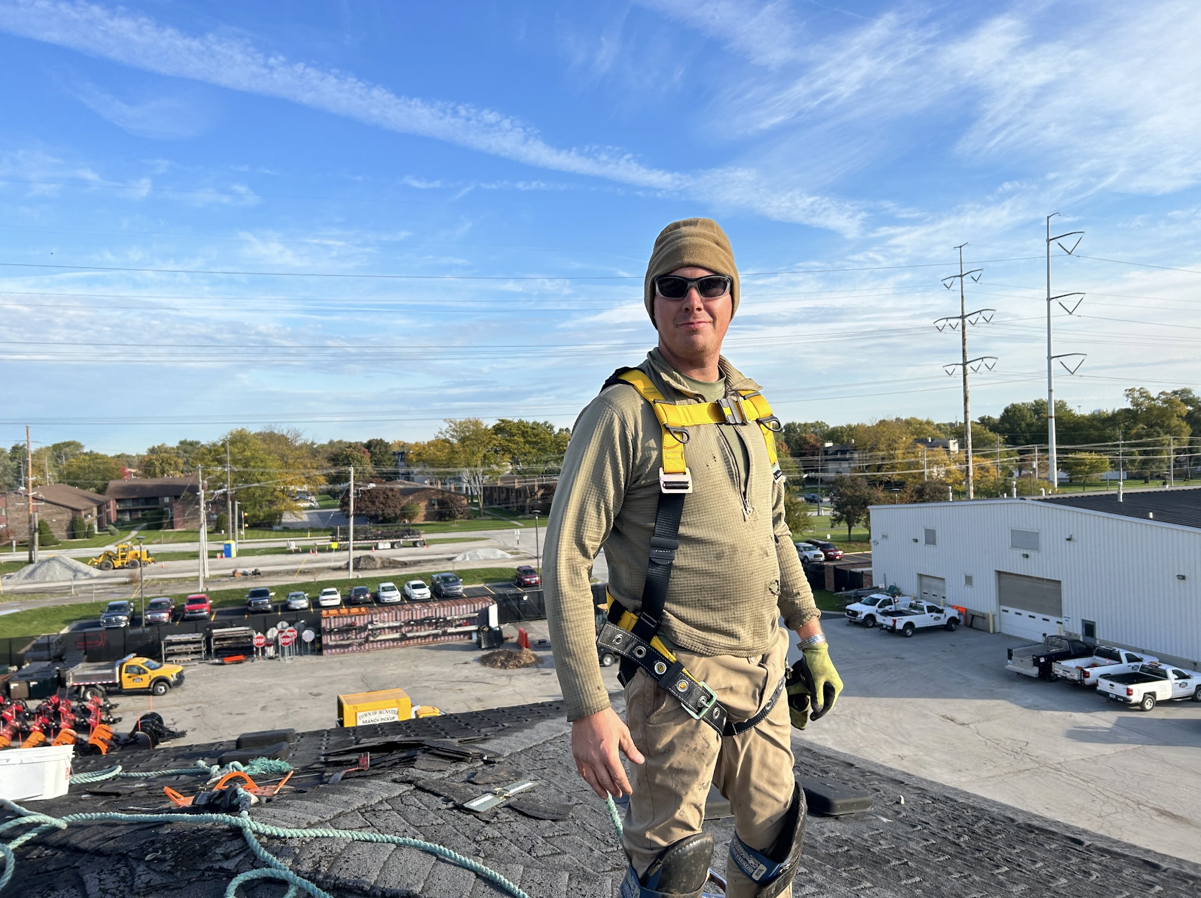 A man wearing protective sunglasses, a tan beanie, gloves, and a harness stands on a rooftop, with a cityscape and blue sky with wispy clouds in the background.