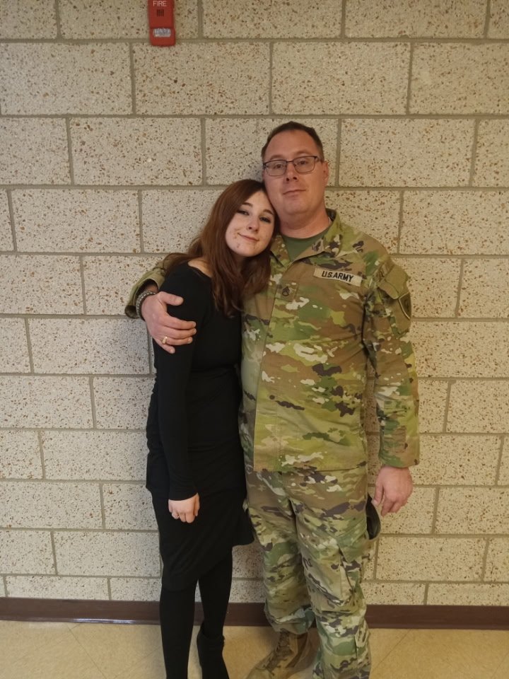 A young woman with long brown hair wearing a black dress and black tights hugging a tall man in military camouflage uniform, standing against a beige brick wall.