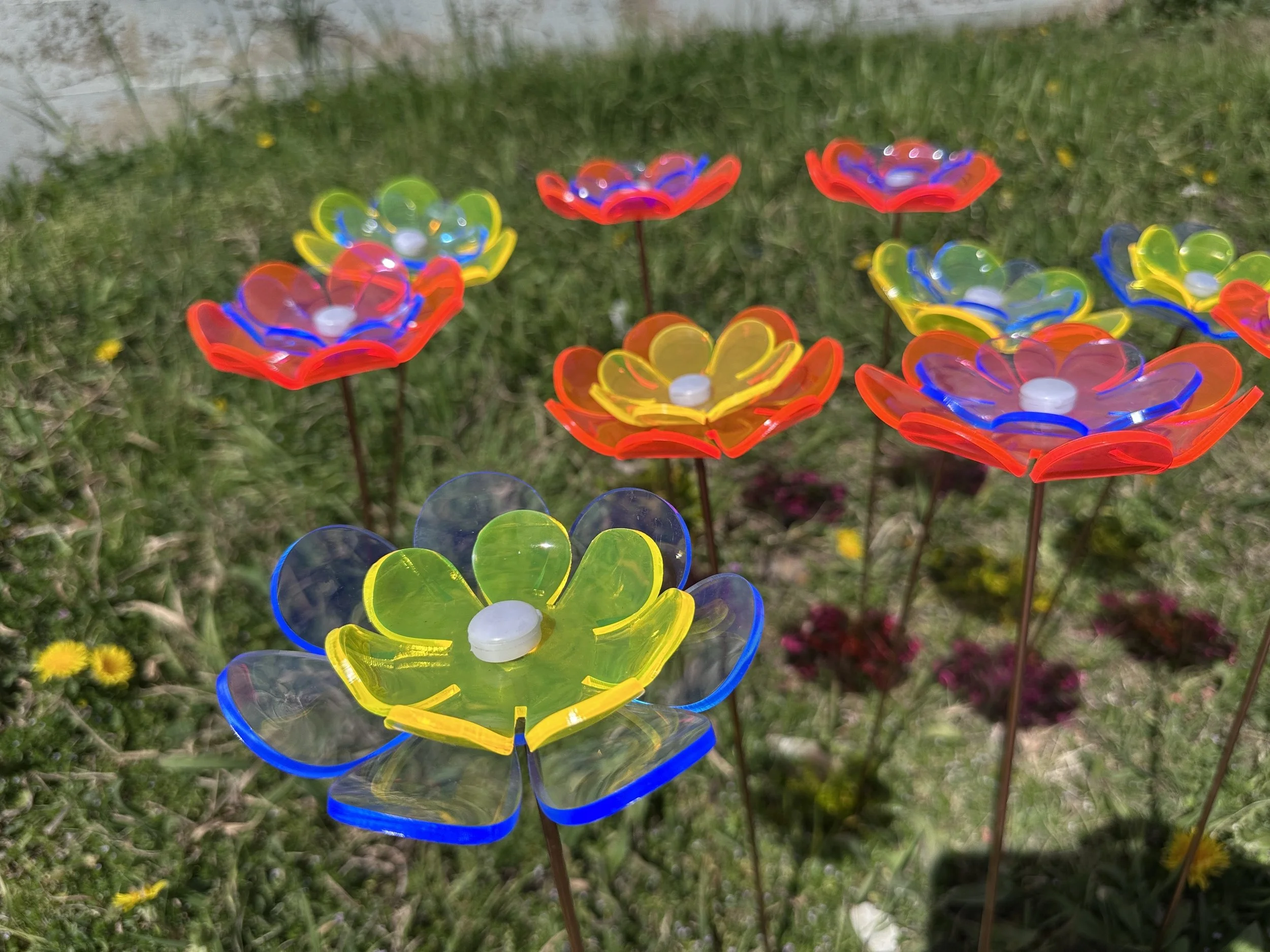 Colorful flower sculptures on metal stems in a grassy field with yellow and pink flowers.