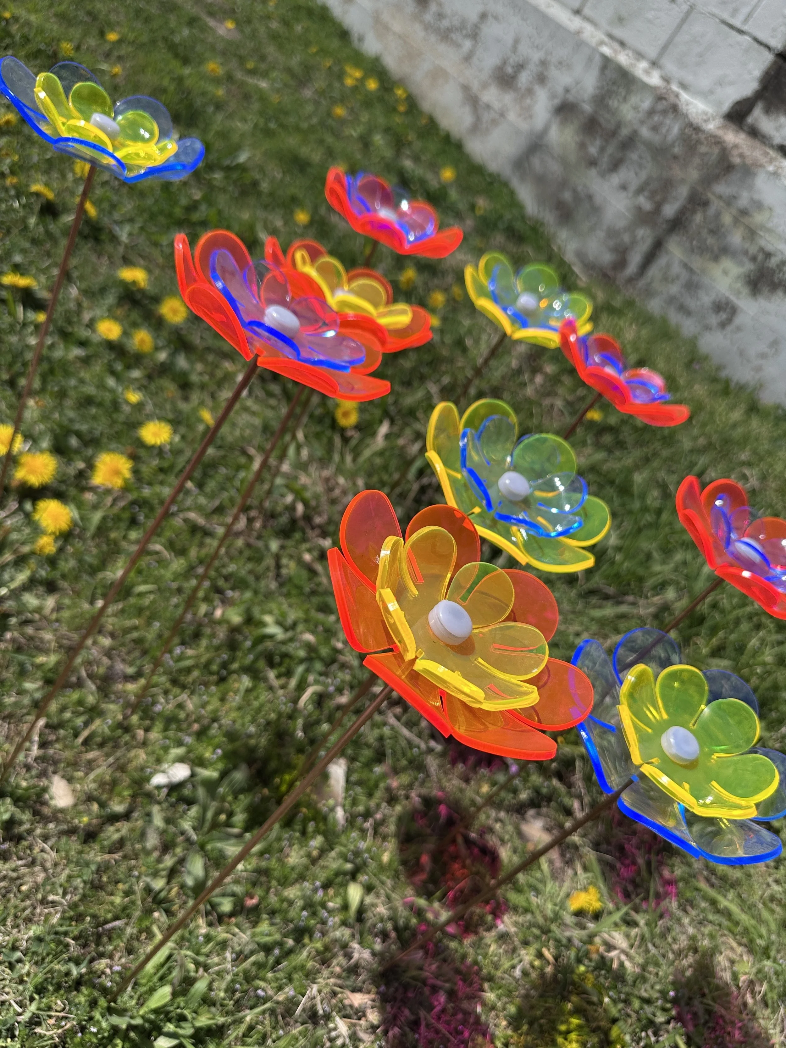 Colorful flower decorations on metal stakes in a grassy area with yellow dandelions and a concrete wall in the background.