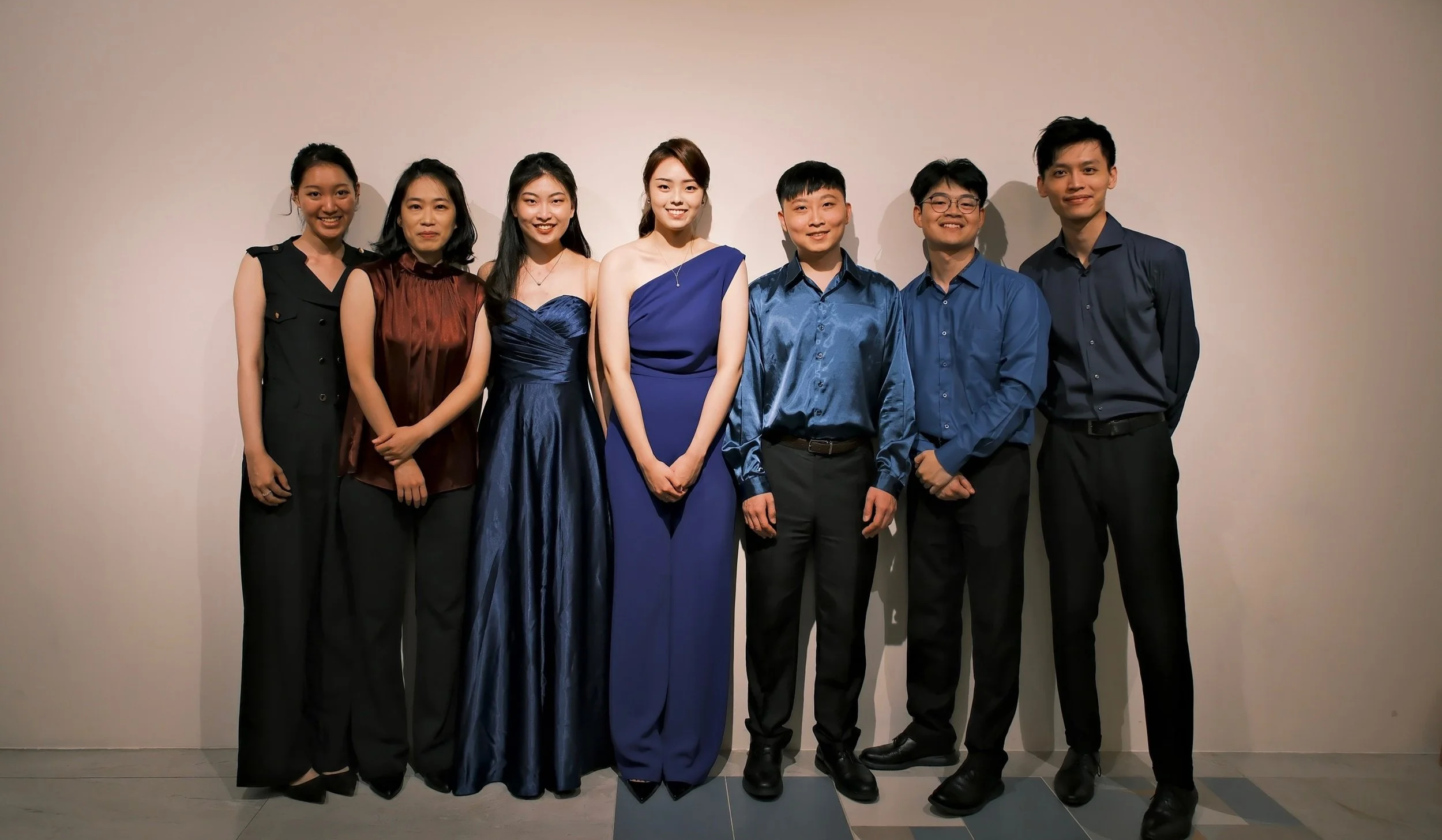 Group of seven young adults in formal attire standing against a plain wall, smiling for the camera.