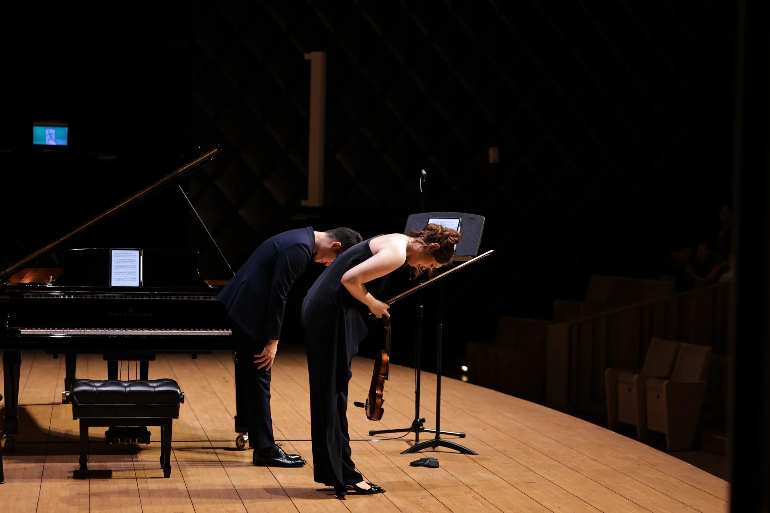 Two performers bowing on a stage after a musical performance, with a grand piano and a cello visible nearby.