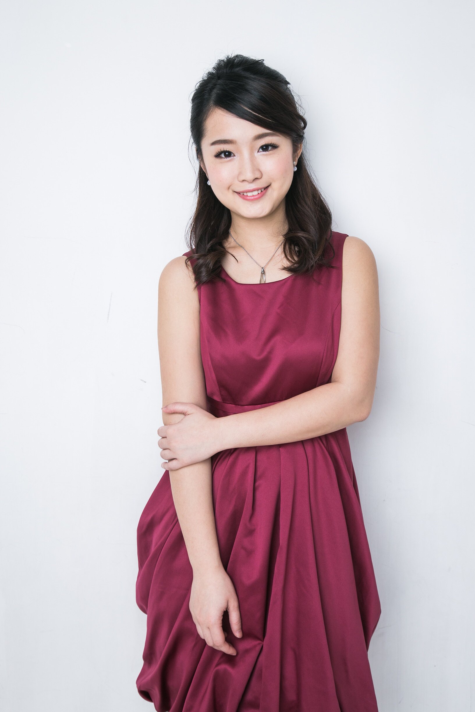 A young woman with dark brown hair styled in loose waves, wearing a sleeveless burgundy dress, diamond earrings, and a pendant necklace, standing against a plain white background.