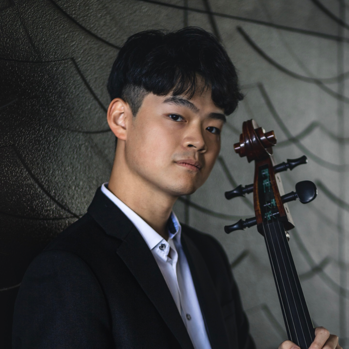 Young man in formal attire holding a cello, standing against a textured wall with abstract lines.