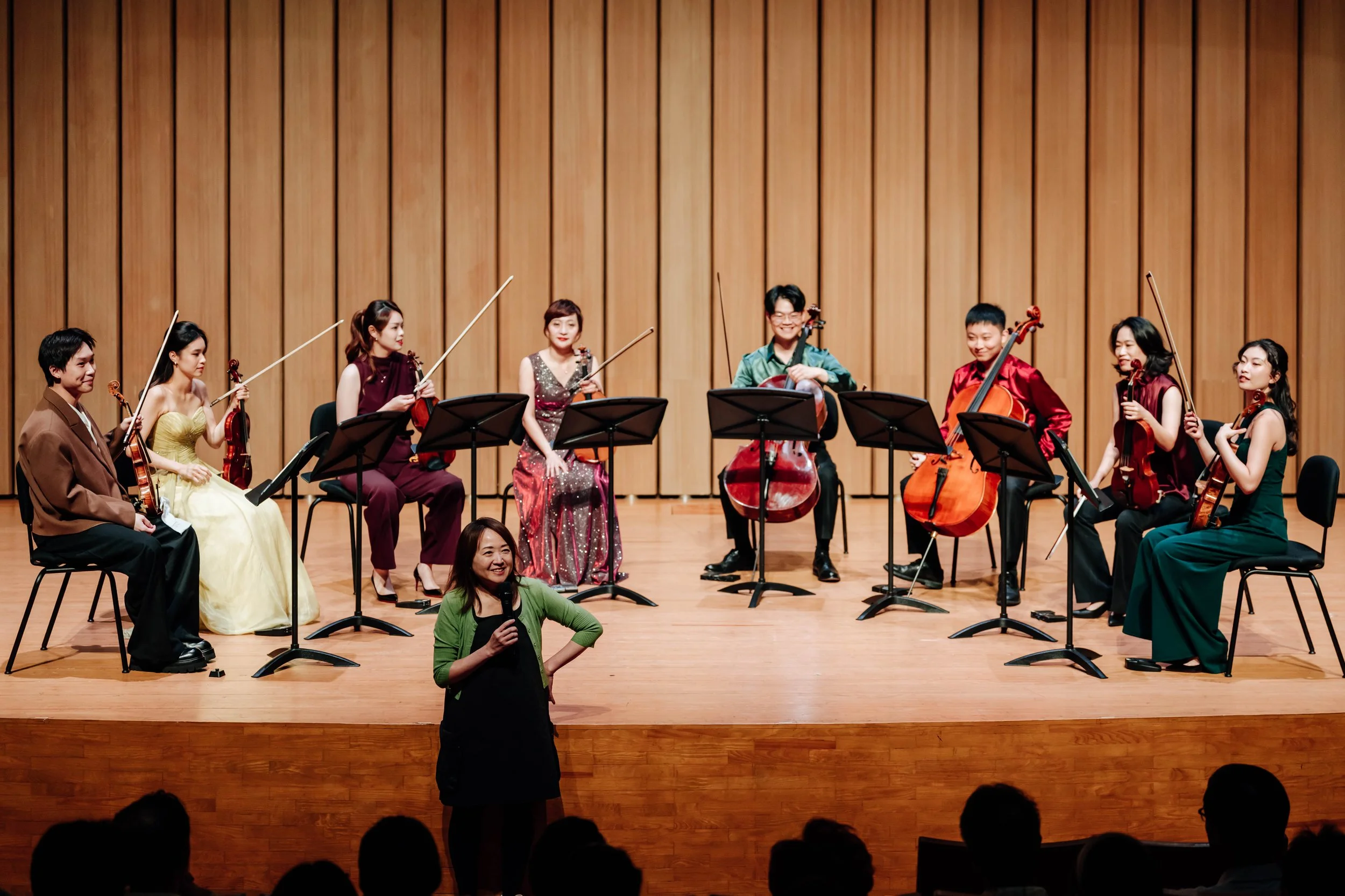 Orchestra with musicians holding violins and cellos on a stage with a wooden backdrop. A woman in a black dress is standing in front of the stage, smiling and talking to the audience. Audience members seen at the bottom.