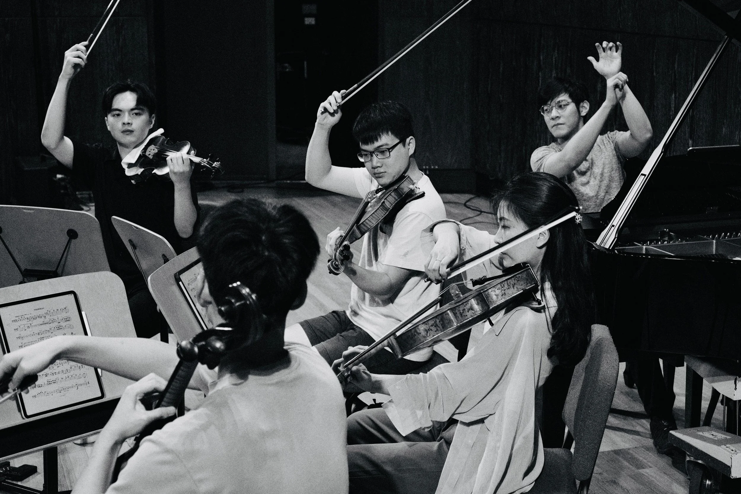 Musicians playing violins and a grand piano during an orchestra rehearsal in a dimly lit music studio.