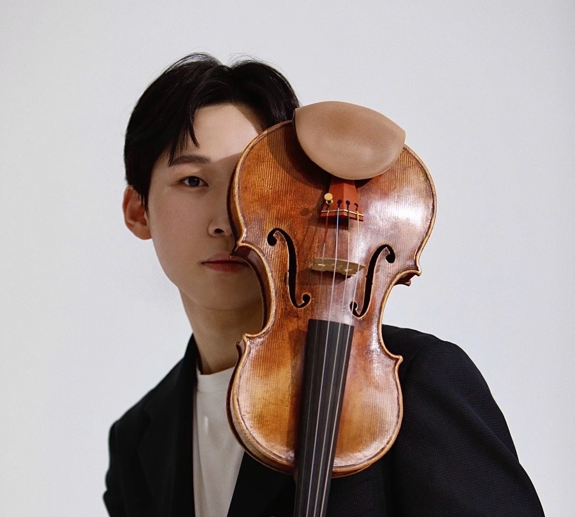 A man holding a violin vertically in front of his face, partially covering it, against a plain white background.