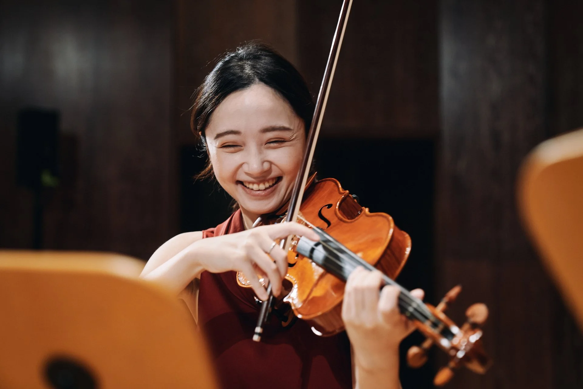 A woman playing the violin, smiling joyfully, in a music studio or concert hall, with a blurred background and a focus on her face and instrument.