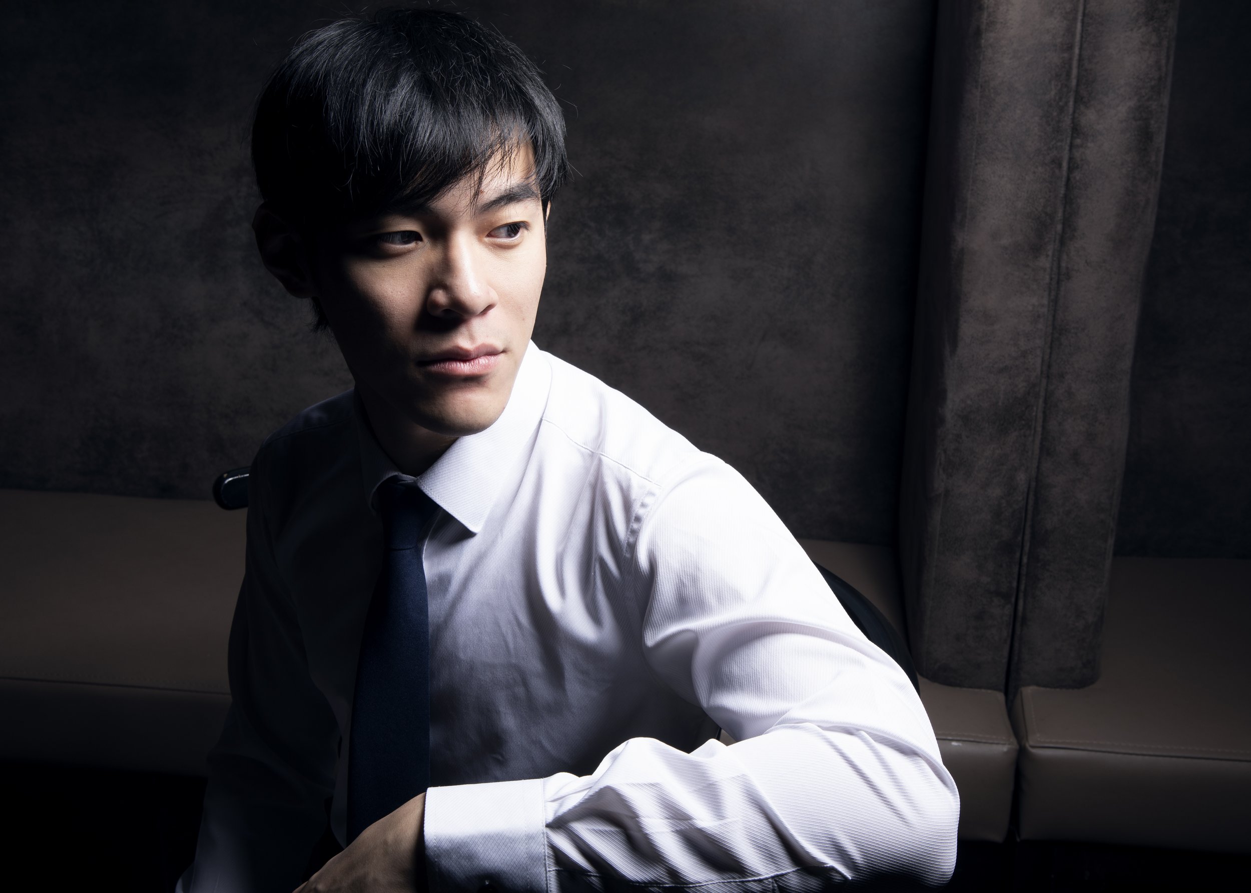 A young man with black hair and fair skin sitting in the backseat of a car, wearing a white shirt and black tie, looking to the side with a serious expression. The car interior is dark, with a textured dark wall in the background.