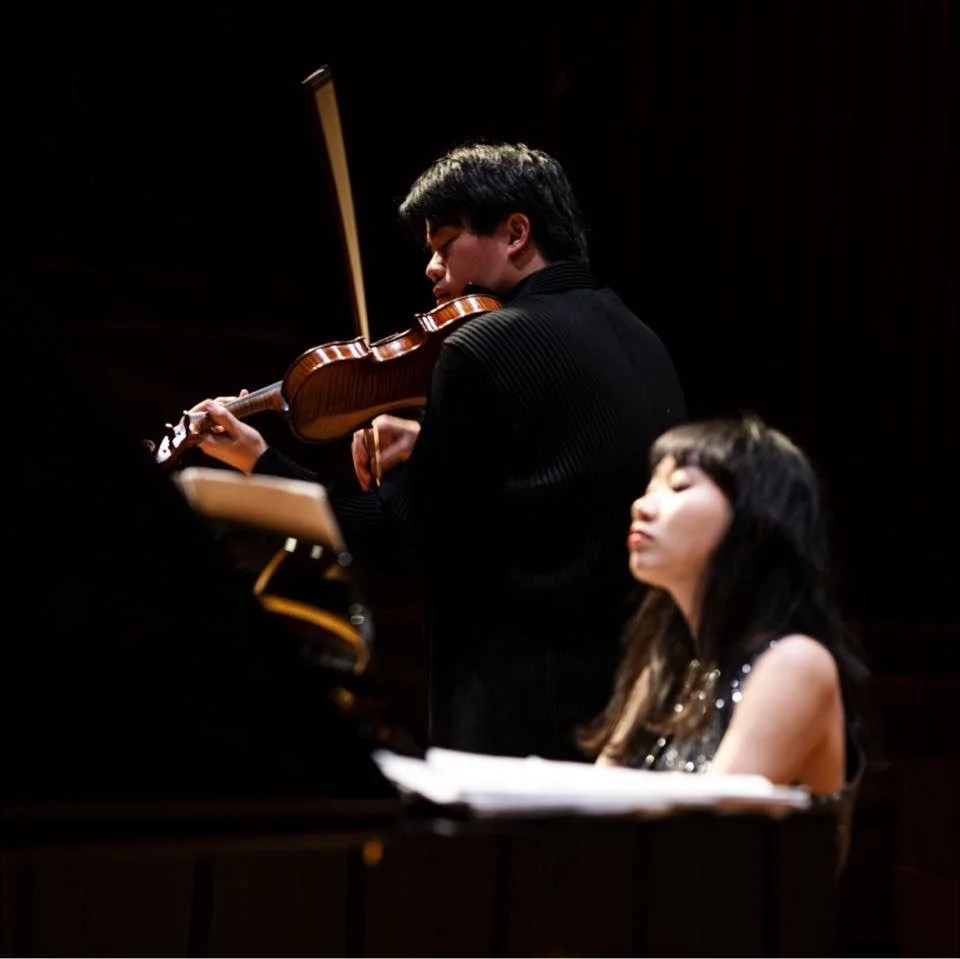 A young man playing a violin on stage with a woman sitting nearby, possibly a pianist, in a dark concert hall.