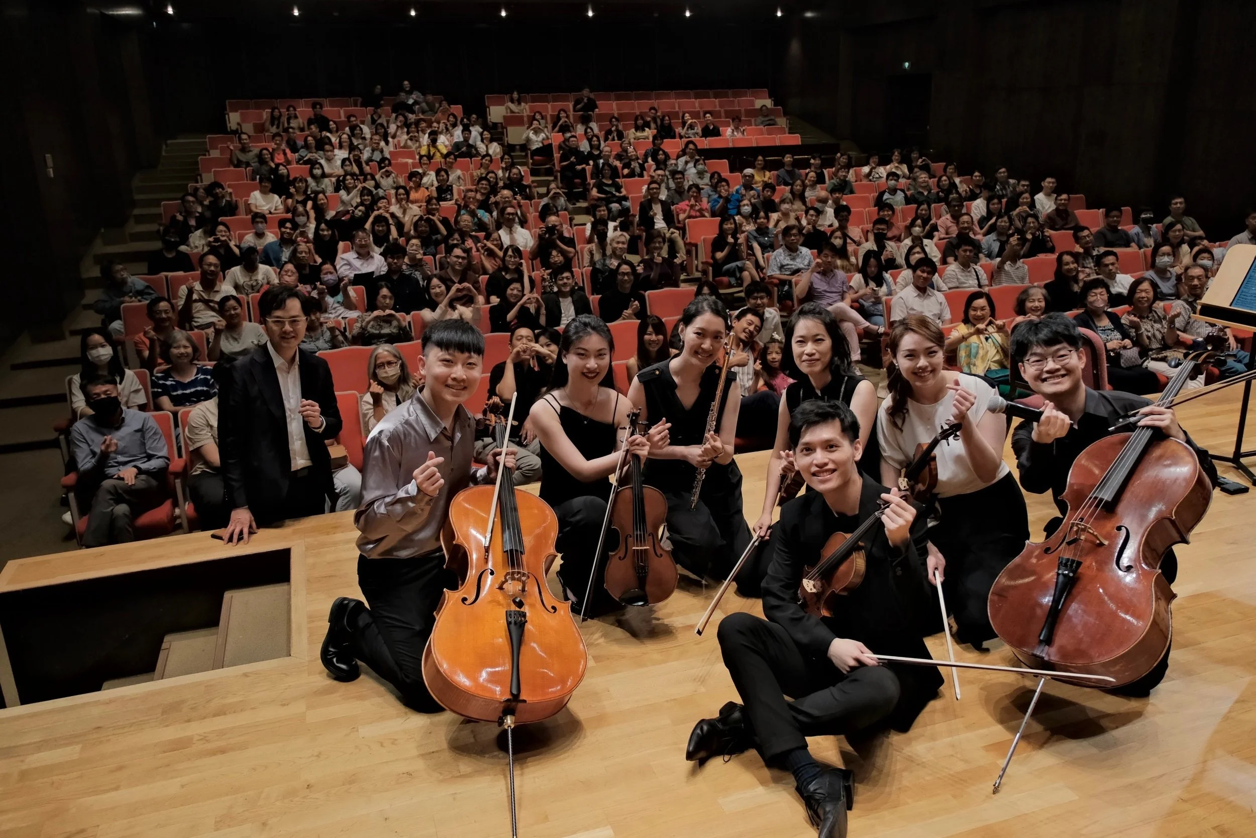 A large audience seated in a concert hall watching performers with musical instruments, including cellos and violins, posing for a photo.