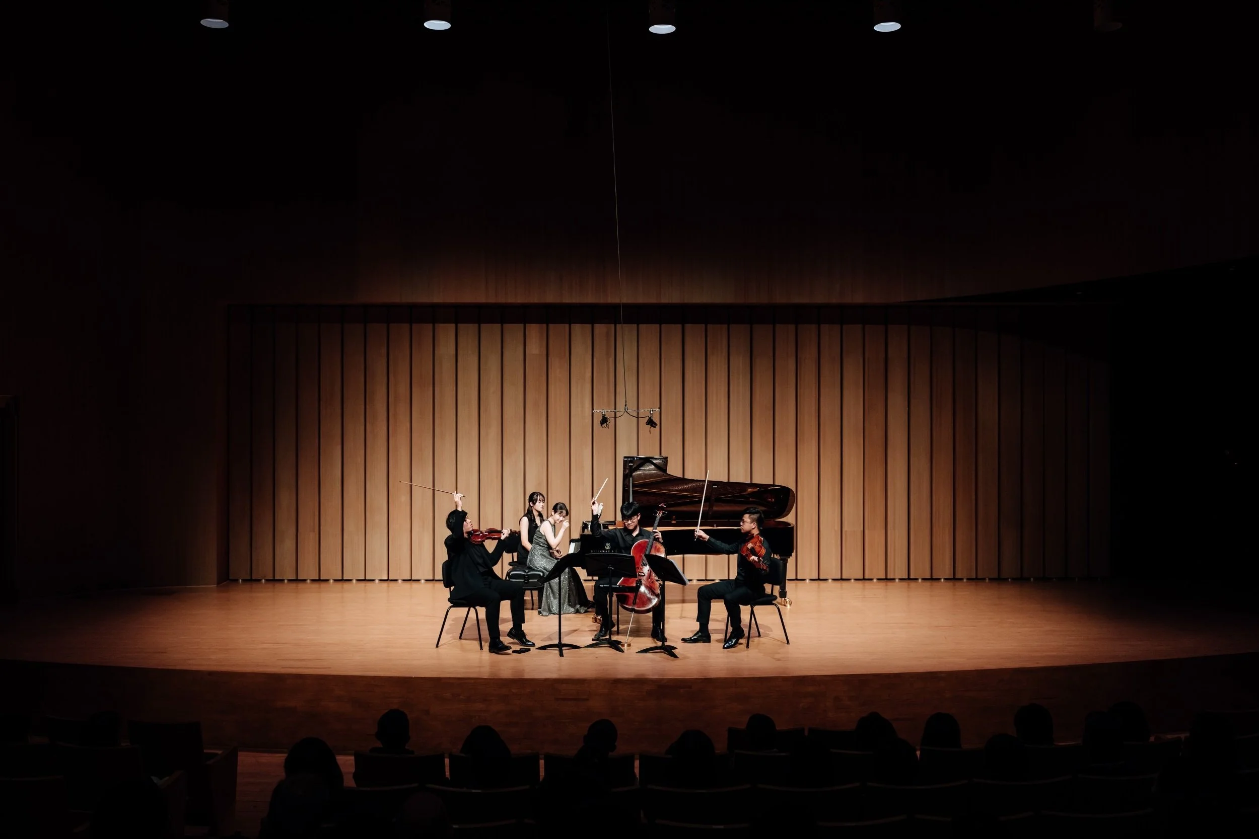 A classical music performance on stage with six musicians, including a pianist, violinists, and a cellist, performing in a concert hall with wooden paneling and stage lighting.