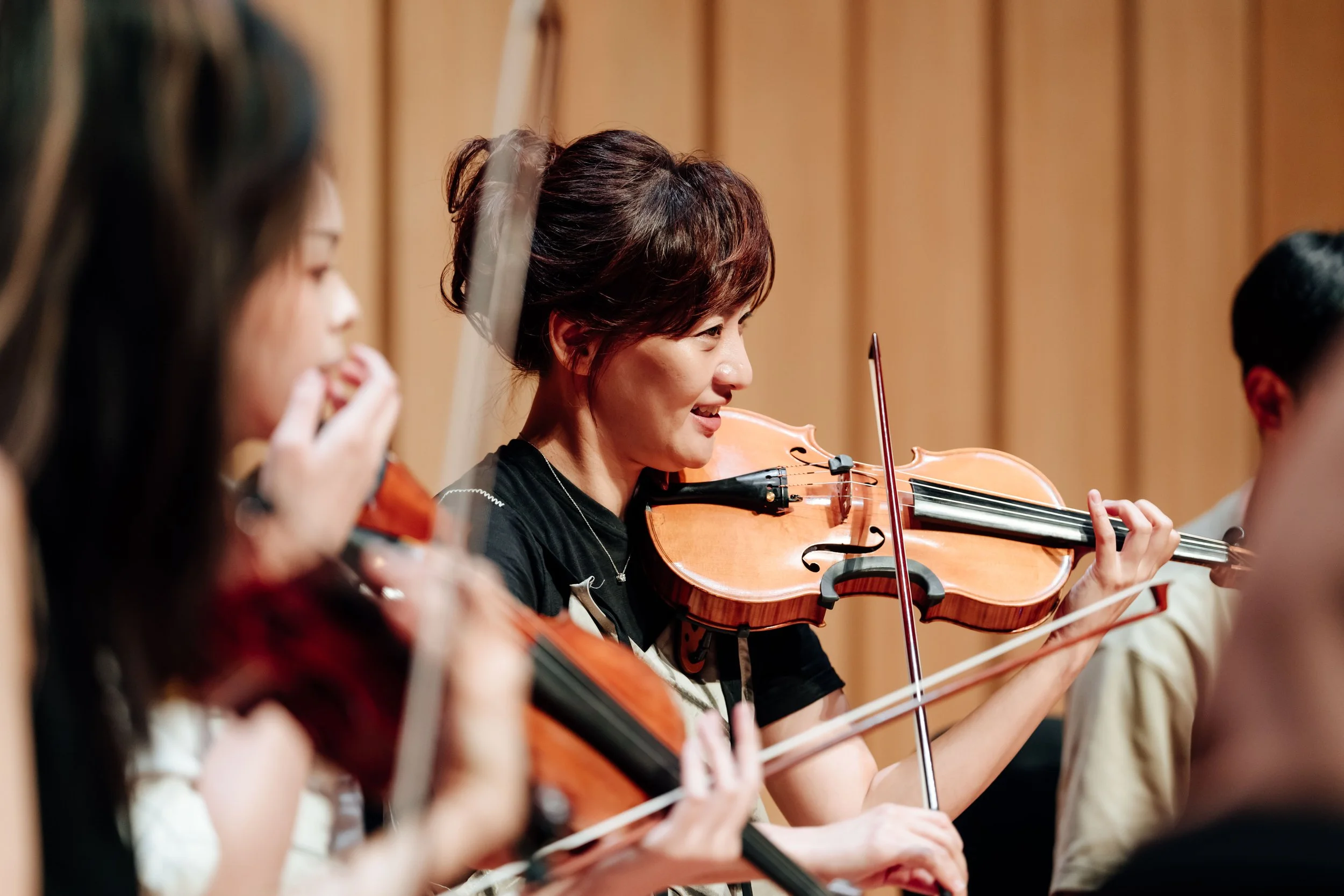 A group of women playing violins in an orchestra, focusing on a woman with short brown hair smiling while playing her violin, with a wooden stage background.