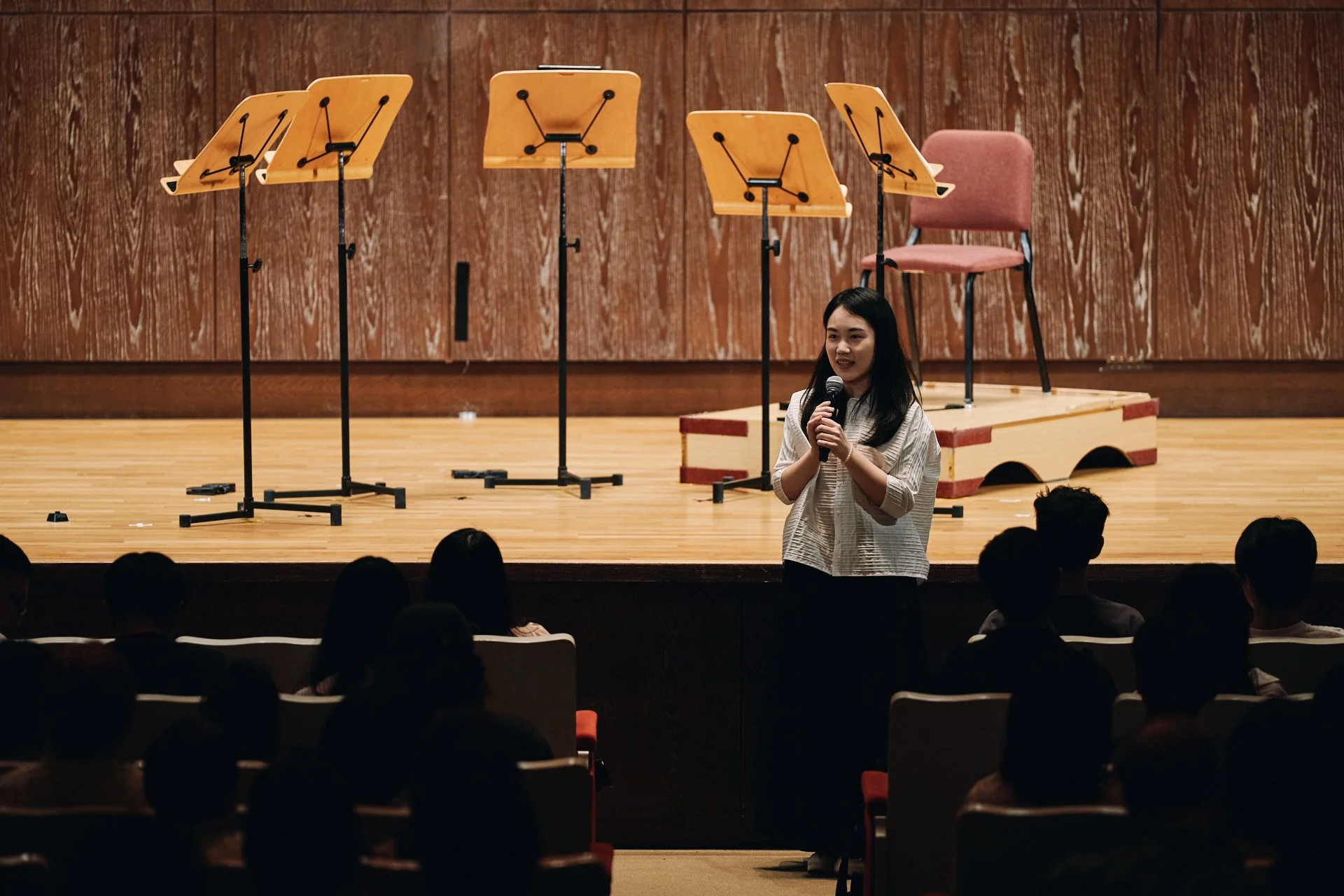 A young woman with long black hair positioned on stage in front of an audience, holding a microphone, with five empty music stands behind her, and a wooden chair on a platform against a wooden-paneled wall.