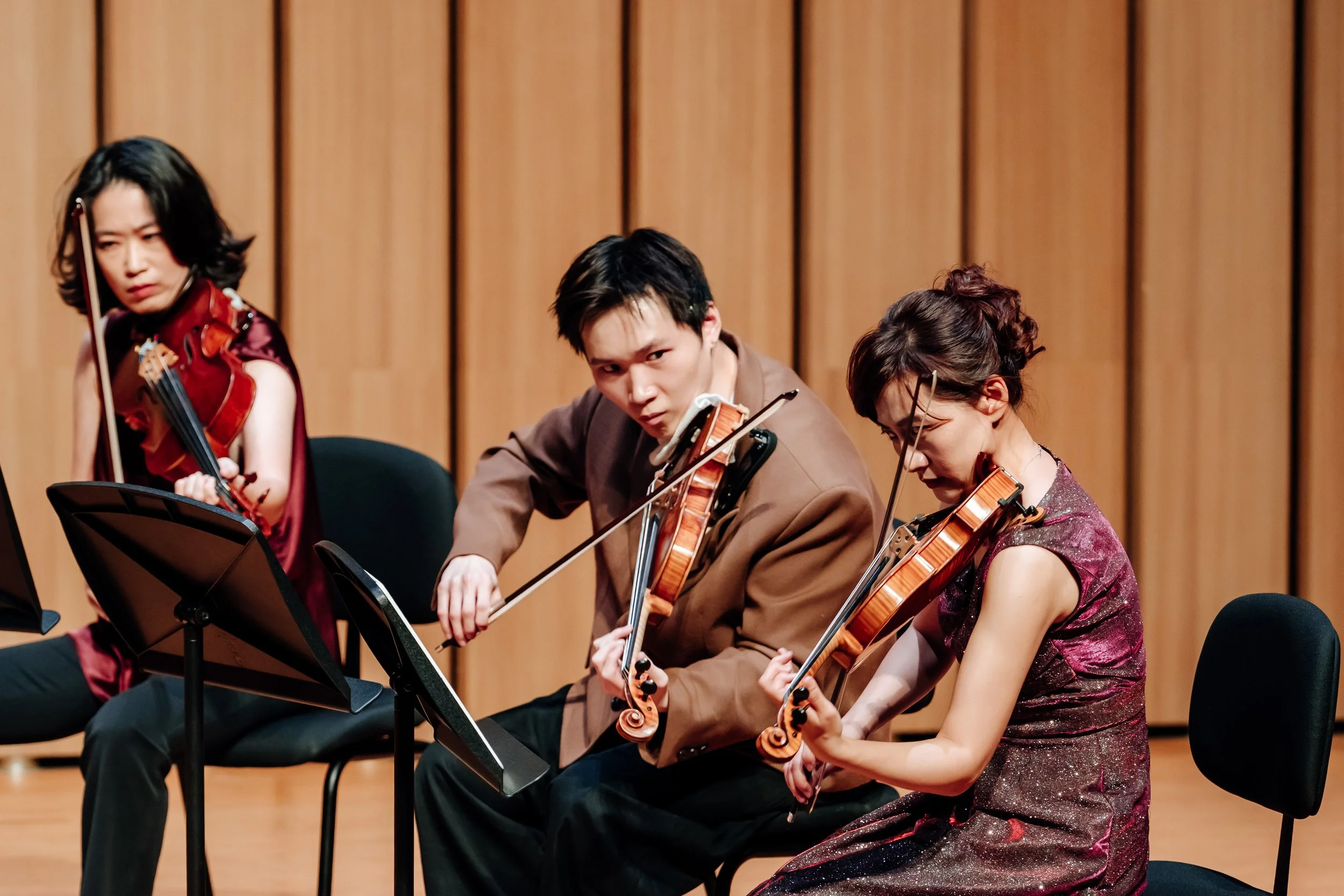 Orchestra musicians playing violins on stage against wooden panel background.